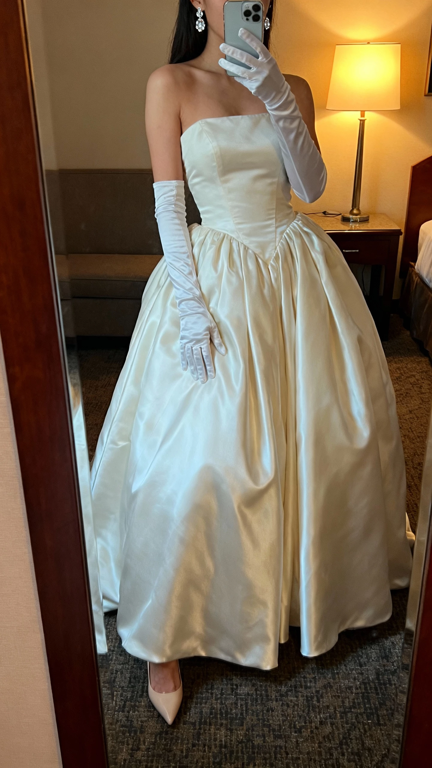 iPhone mirror selfie of a woman wearing long opera gloves with a satin ballgown, subtle crystal earrings, and classic heels peeking, face not visible, cropped at neck, hotel suite full-length mirror, warm evening lamp light, natural iPhone photo quality