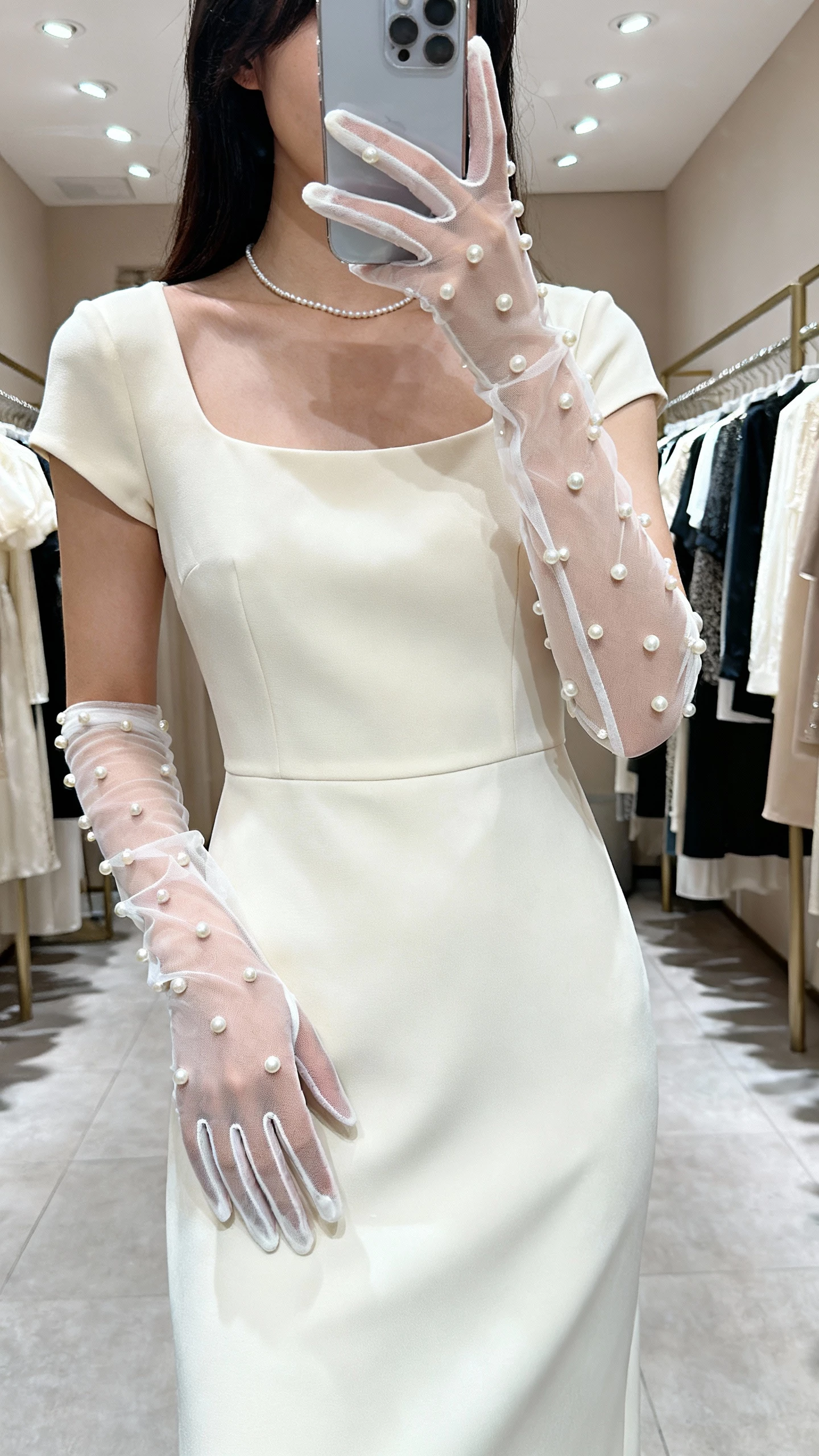 iPhone mirror selfie of a woman wearing pearl-embellished mesh gloves with a classic boatneck dress, thin pearl necklace, face not visible, boutique fitting room, overhead indoor lighting, natural iPhone photo quality
