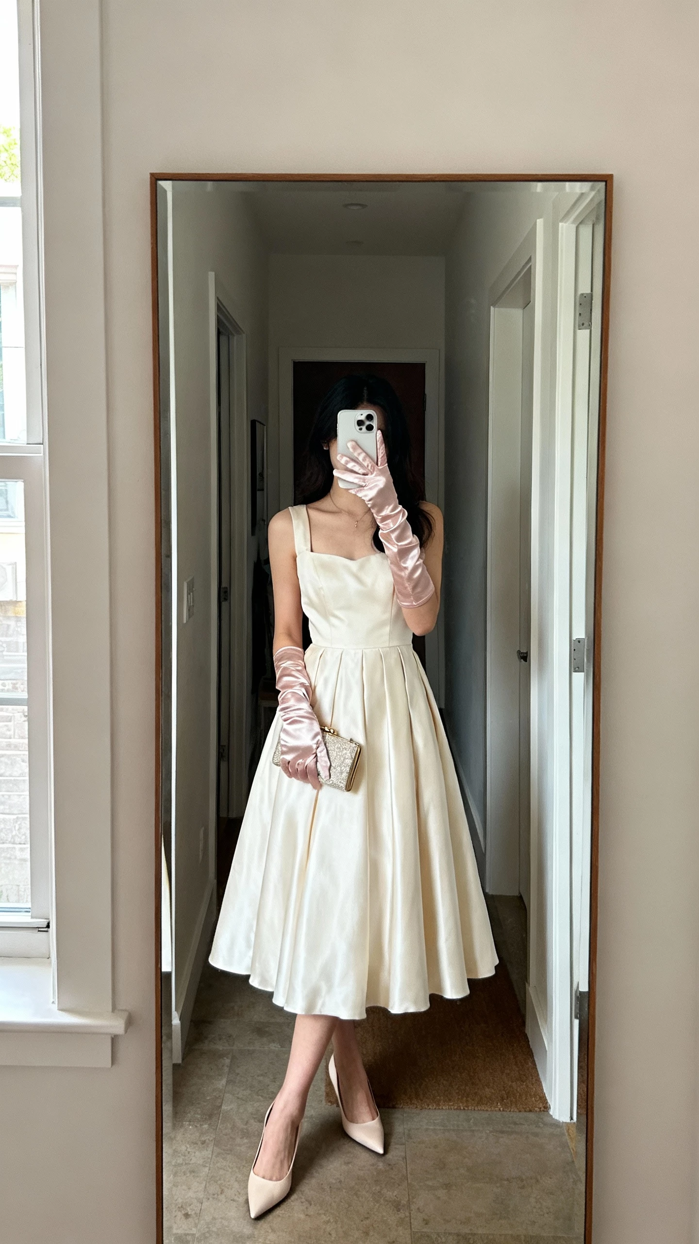 iPhone mirror selfie of a woman wearing satin wrist gloves with a timeless tea-length dress, kitten heels and a small clutch, face not visible, apartment entryway mirror, natural window light, natural iPhone photo quality