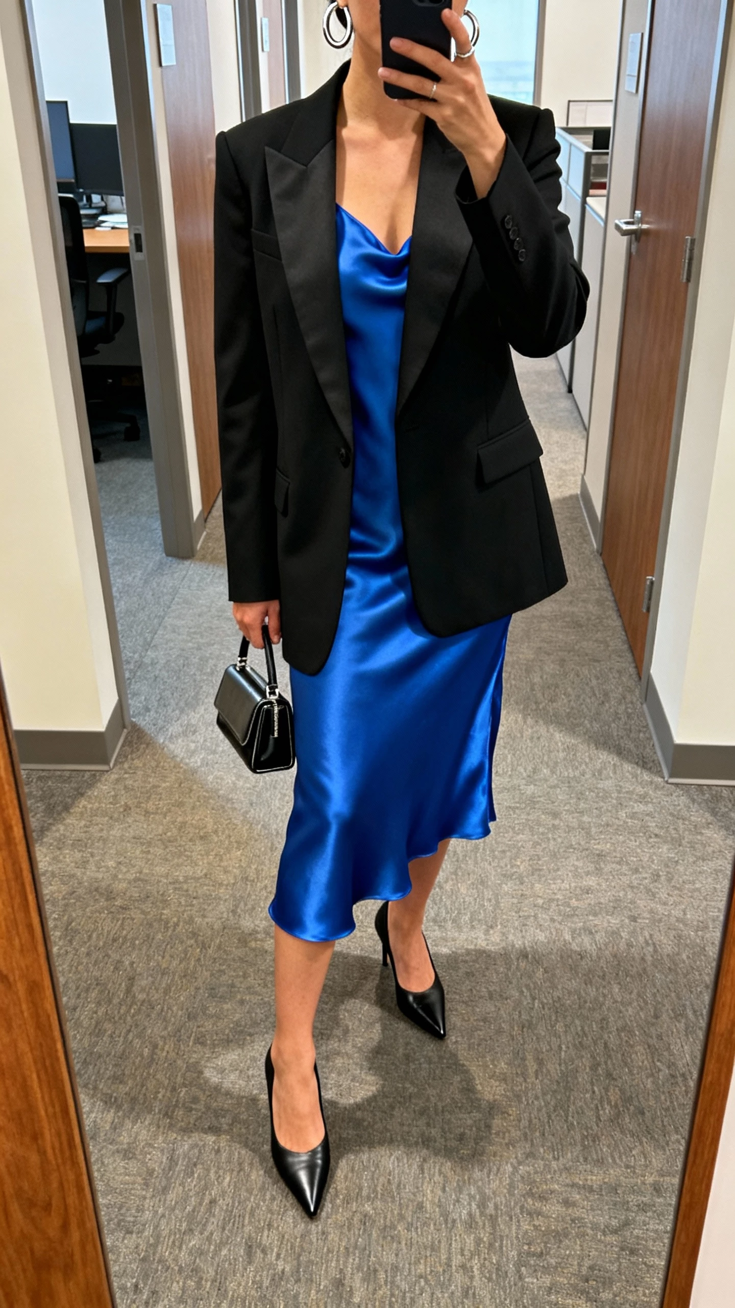 iPhone office hallway mirror selfie of a woman in a sapphire satin slip dress layered under a sharp black tuxedo blazer, pointed black pumps, sleek silver hoops, and a structured mini bag, face not visible, natural indoor lighting, casual iPhone aesthetic.