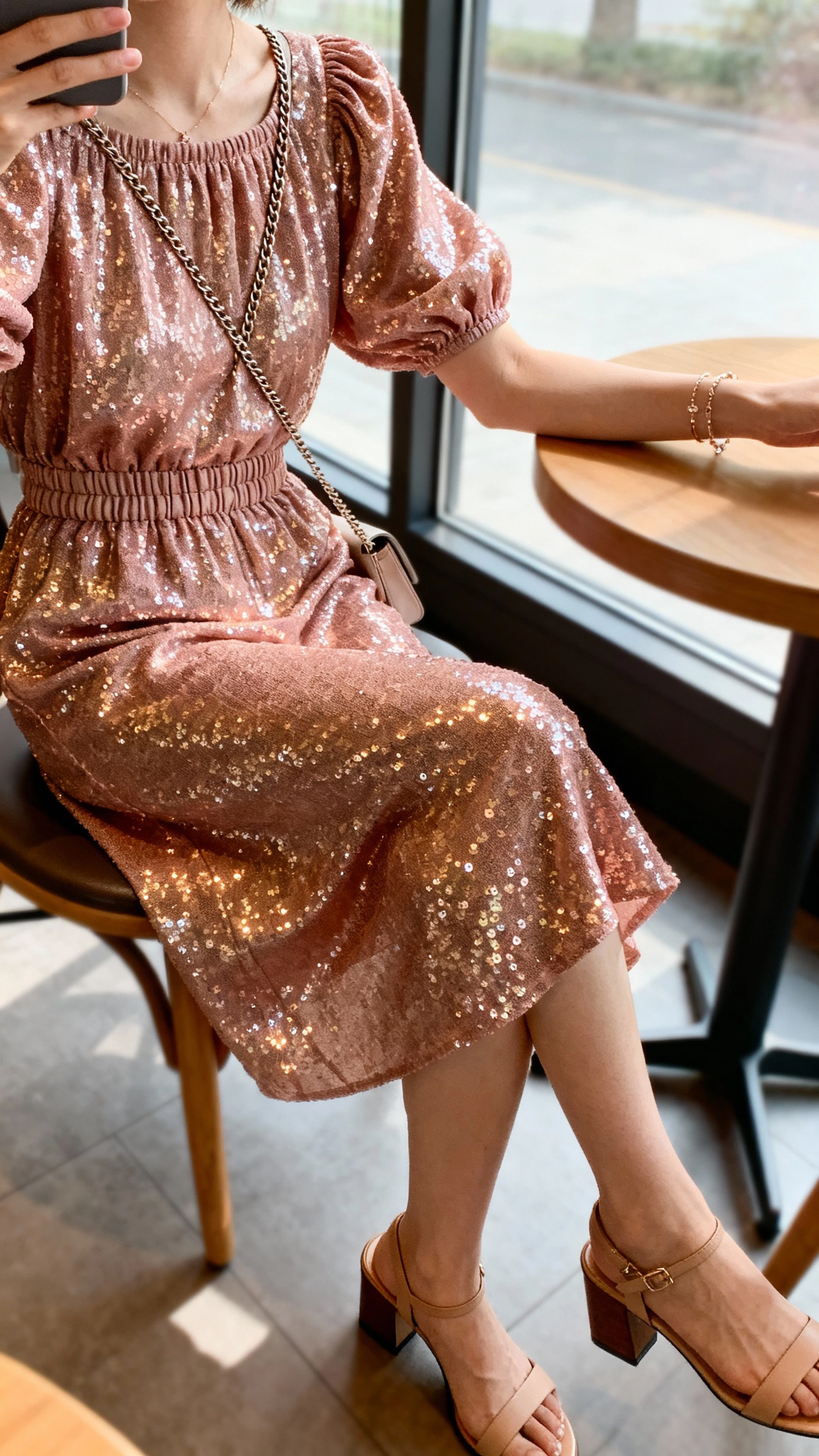 iPhone seated cafe selfie, cropped at neck, of a woman wearing a rose gold sequin dress with a blouson bodice and elastic waist, block-heel sandals, dainty bracelets, and a crossbody chain bag, face not visible, morning window light, casual iPhone aesthetic.