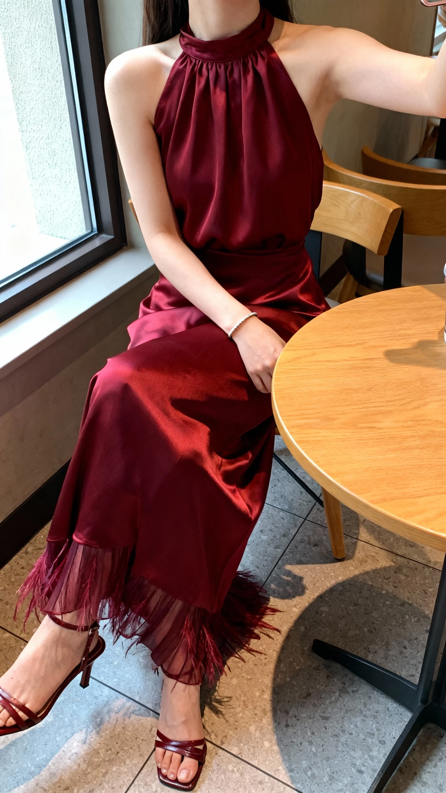 iPhone seated cafe selfie cropped at neck showing a woman in a burgundy satin halter gown with featherlight pleats, simple bracelet, heeled sandals, face not visible, window-side table, morning light, casual iPhone photo.