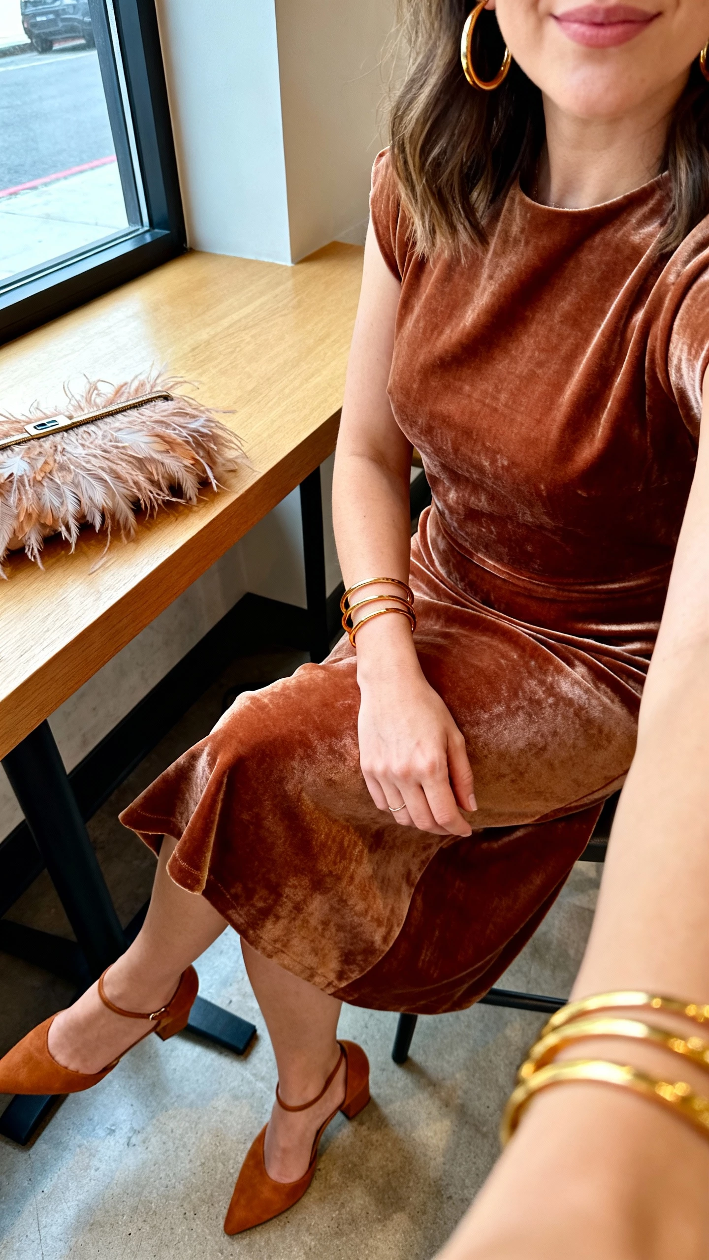 iPhone seated cafe selfie cropped at shoulders of a woman in bronze velvet midi, feathered clutch on the table, warm metal (gold) hoops and bangles, tan heels, face not visible, window-side table, natural daylight, iPhone photo.