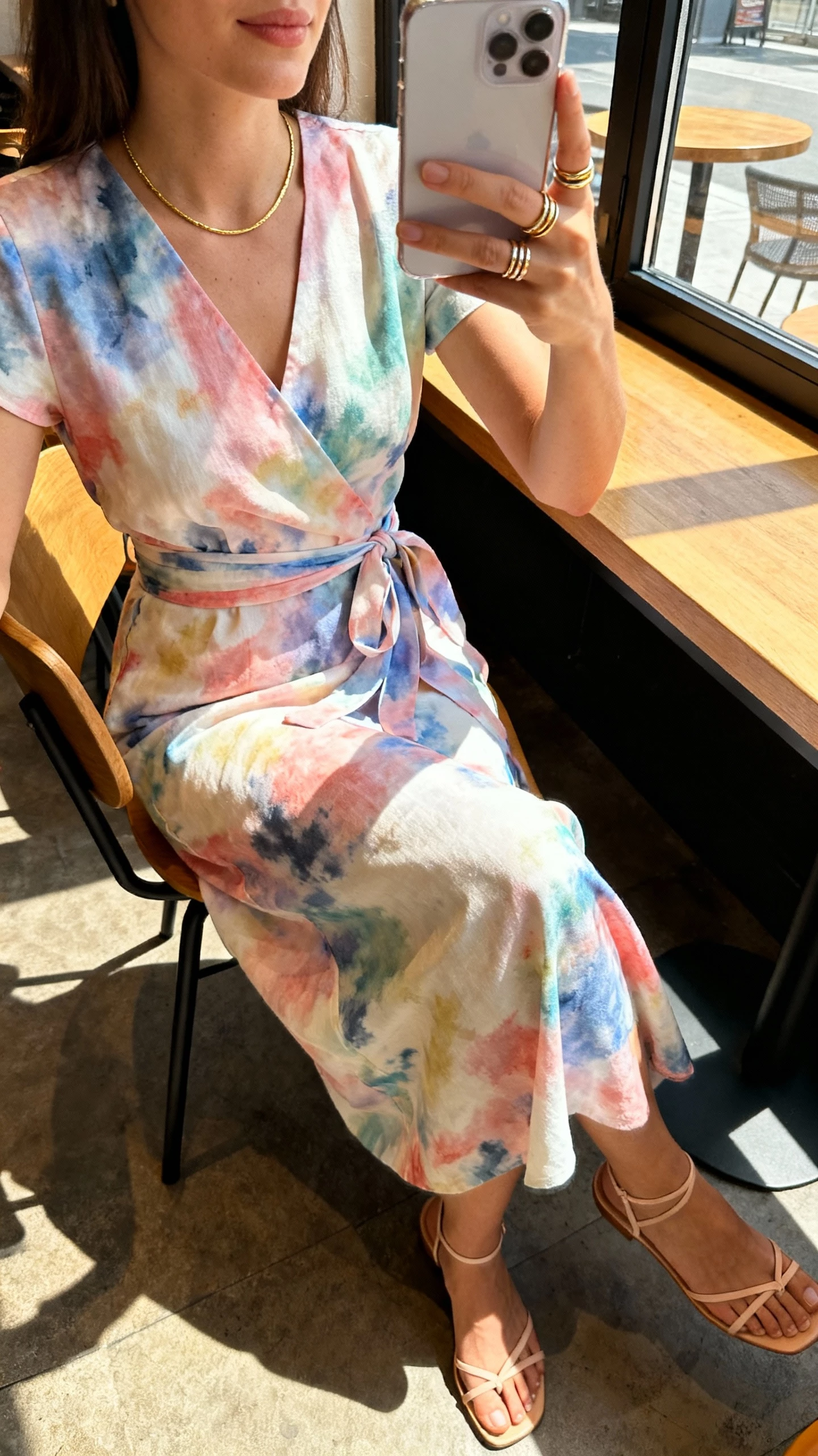 iPhone seated cafe selfie cropped at shoulders showing a woman in a watercolor wrap dress with barely-there sandals, slim gold necklace and stackable rings, face not visible, sunlit cafe window, natural iPhone quality.