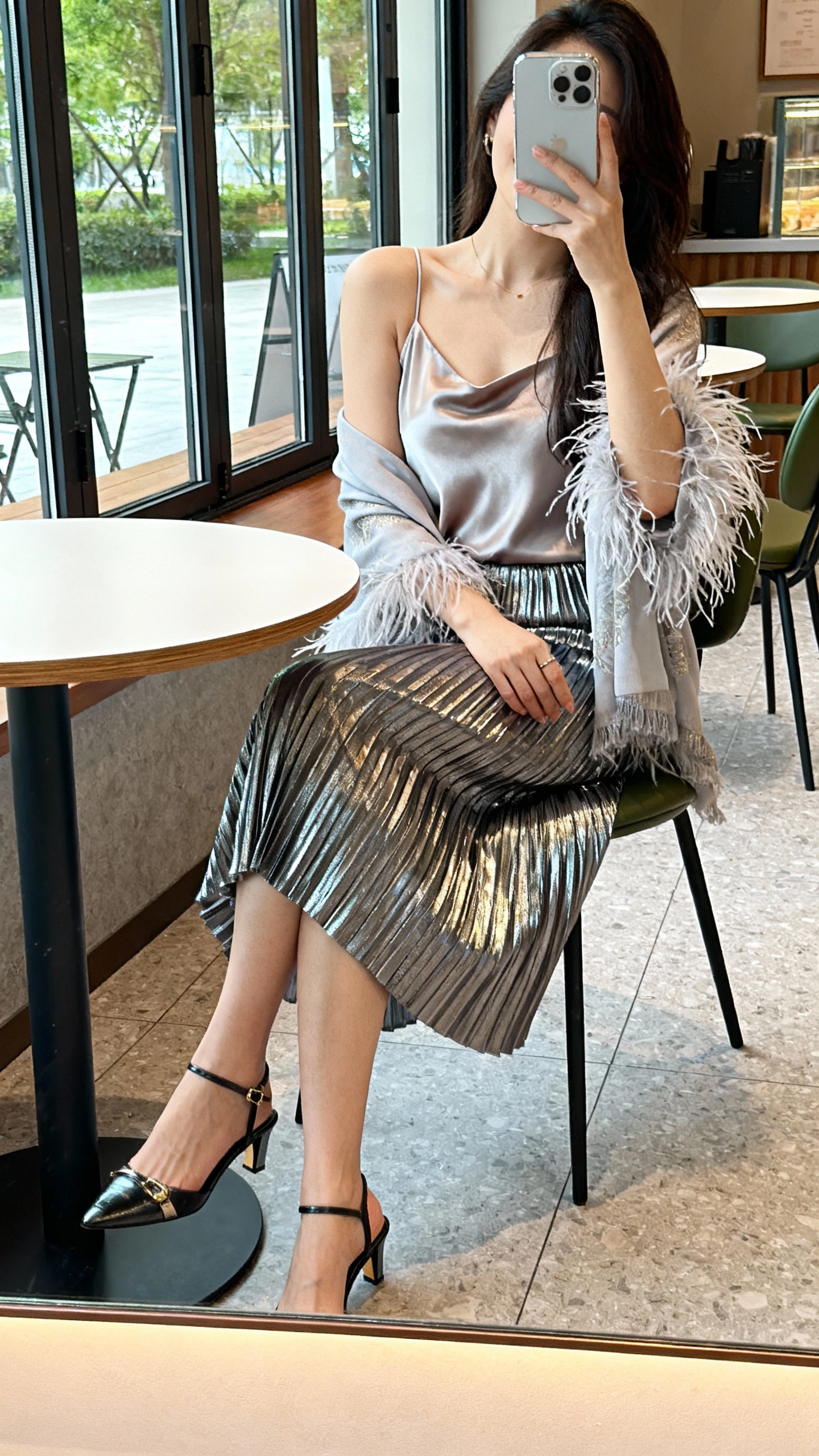 iPhone seated cafe selfie, table reflection capturing a woman in a metallic pleated skirt, silk camisole, and a feather-trim shawl, slingback heels, face not visible, daylight by cafe window, natural iPhone quality.