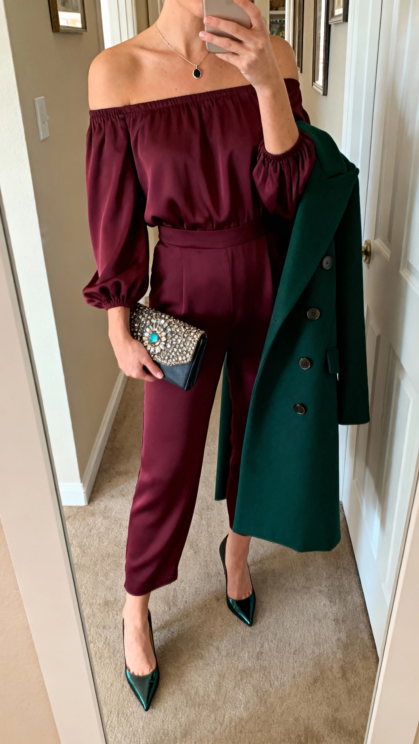 iPhone selfie cropped at neck showing a woman in an off-the-shoulder wine crepe jumpsuit with statement sleeves, short pendant necklace, satin pointed-toe pumps, gemstone-clasp clutch, deep green double-breasted tailored coat; face not visible, apartment hallway mirror, warm indoor light, natural iPhone aesthetic.