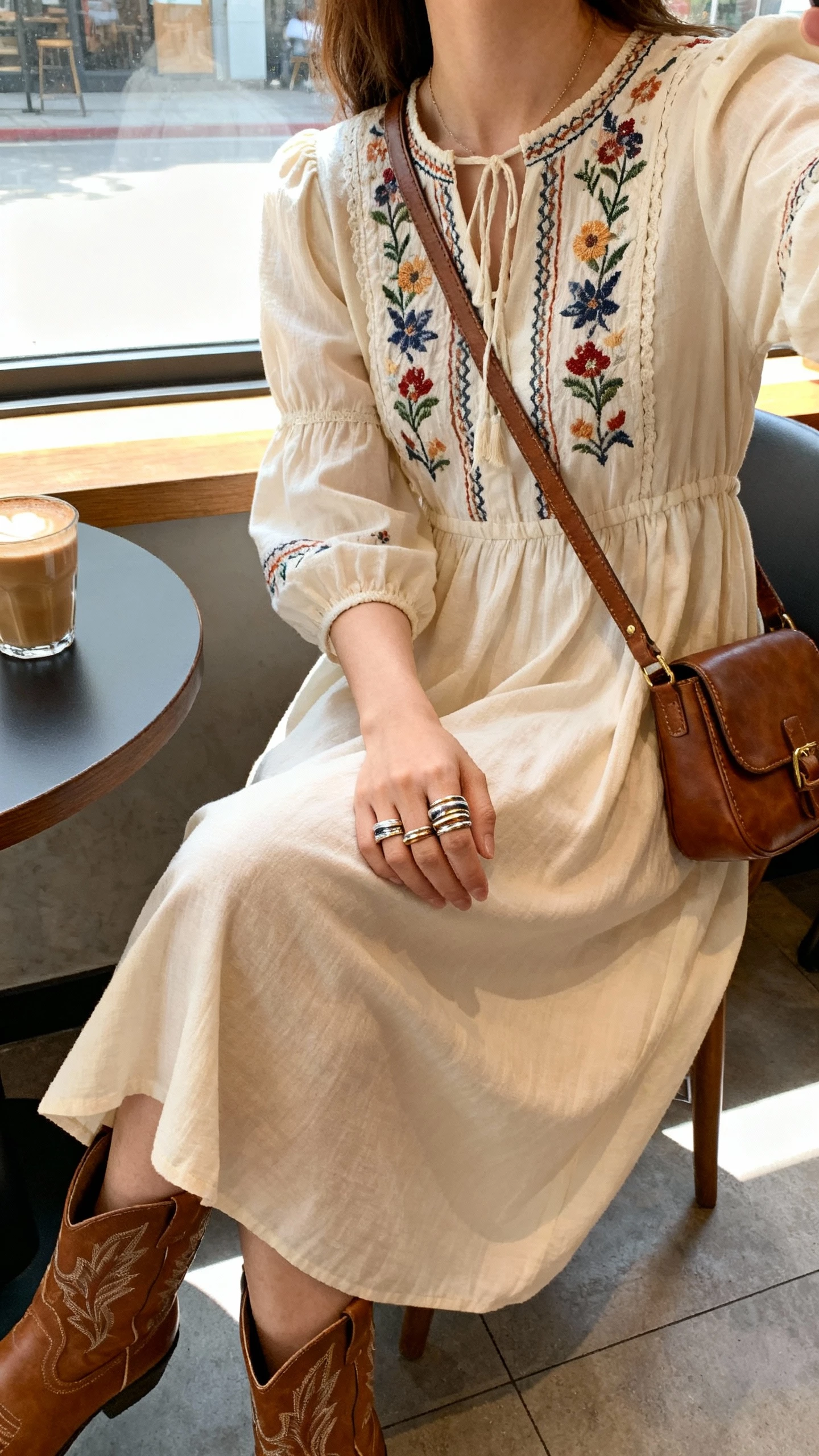 iPhone selfie cropped at neck showing a woman in an embroidered prairie-chic midi dress, western-inspired ankle boots, stacked rings, leather crossbody, face not visible, coffee shop window seat with soft daylight, natural iPhone aesthetic.