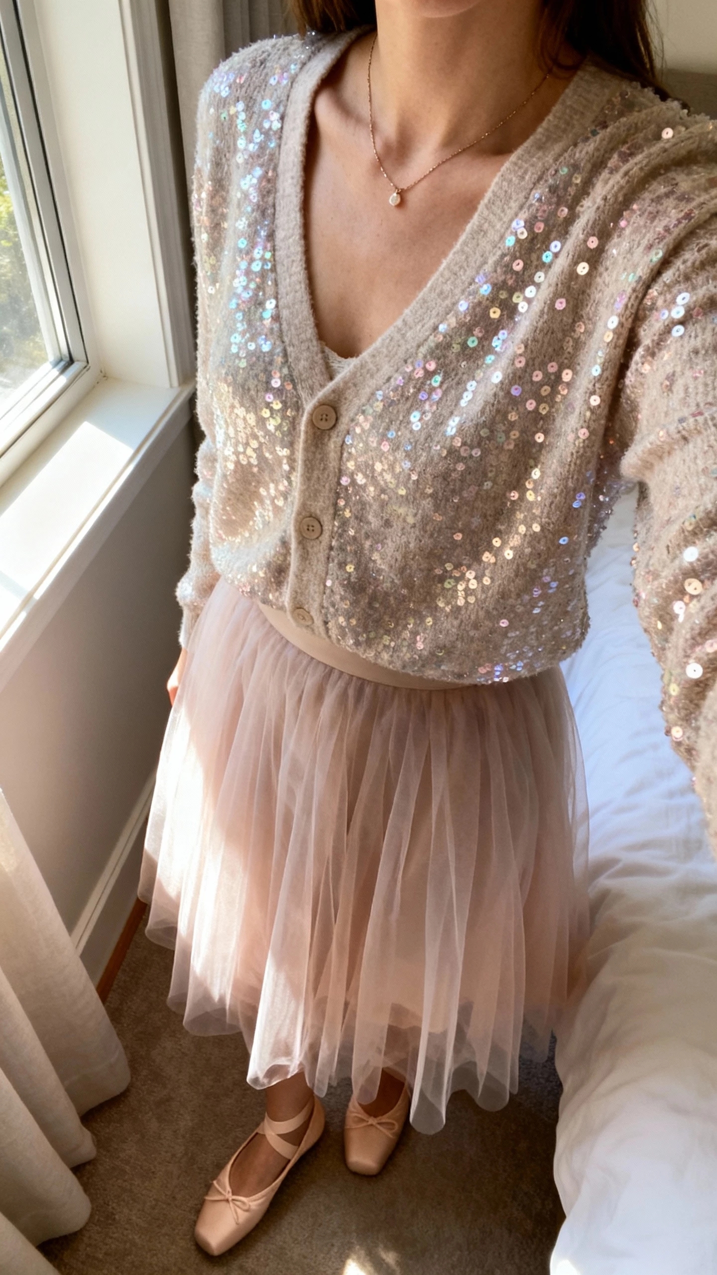 iPhone selfie cropped at neck showing a woman in a soft tulle skirt with a sequin cardigan, dainty necklace, ballet flats, face not visible, cozy bedroom corner, window light, casual iPhone photo.
