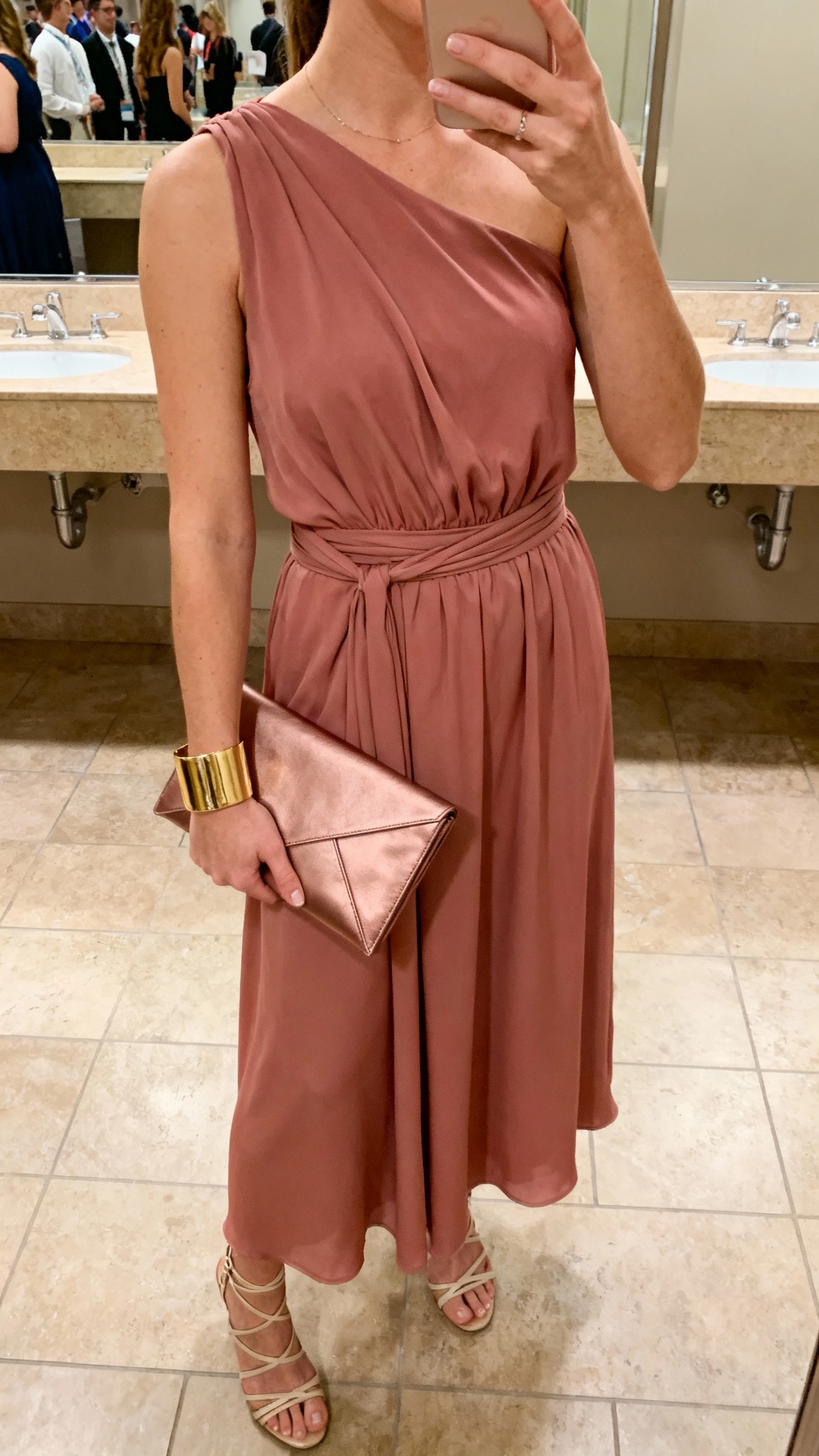 iPhone selfie cropped at neck showing a woman in a dusty-rose one-shoulder dress with gathered waist, gold cuff bracelet, strappy heels, and a sleek envelope clutch, face not visible, bathroom mirror at an event venue, warm indoor lighting, natural iPhone aesthetic.
