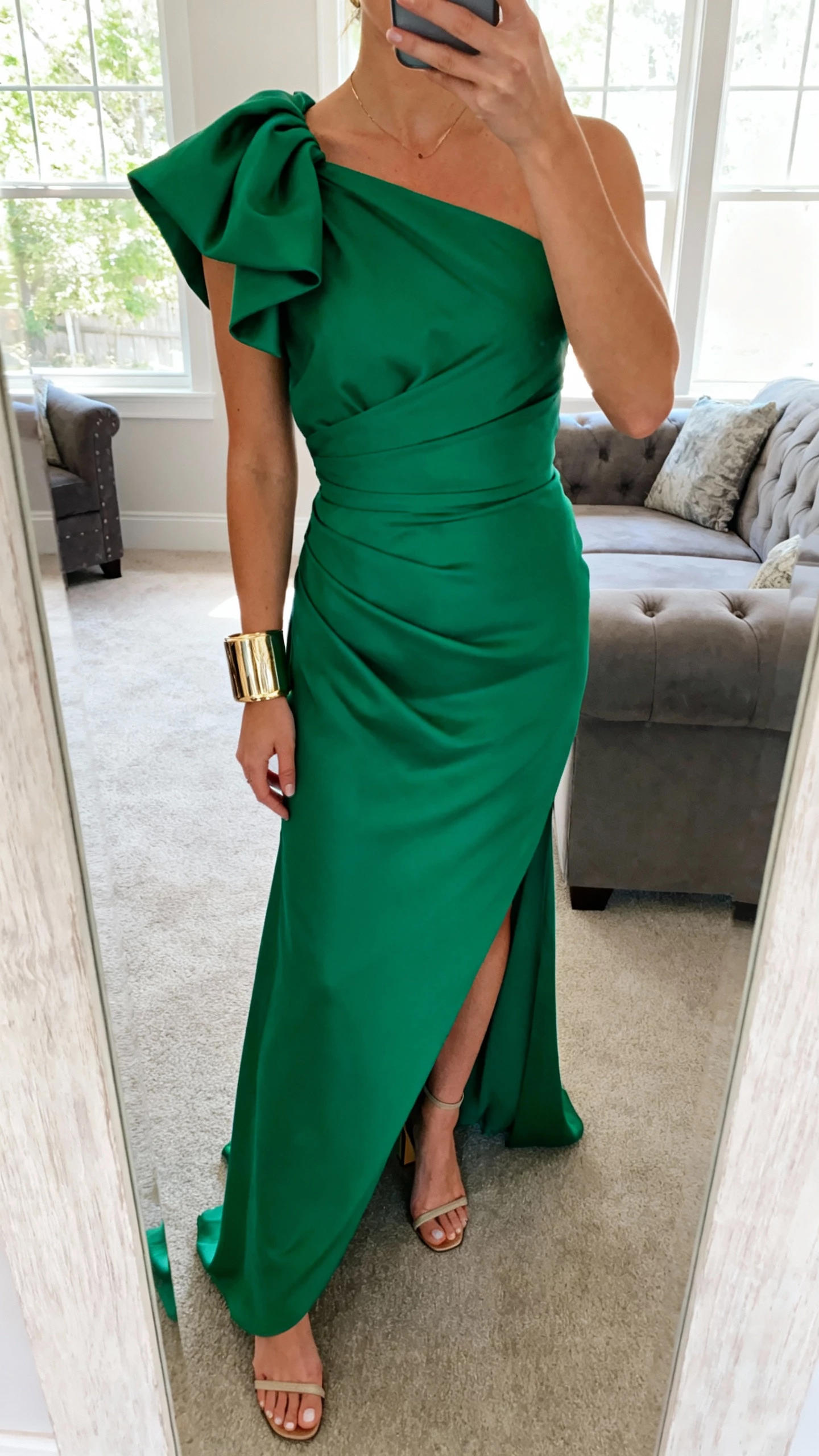 iPhone selfie cropped at neck showing a woman wearing an emerald one-shoulder gown with a sculpted bow at the shoulder, statement cuff bracelet, heeled sandals, face not visible, living room mirror, daylight from window, natural iPhone photo.