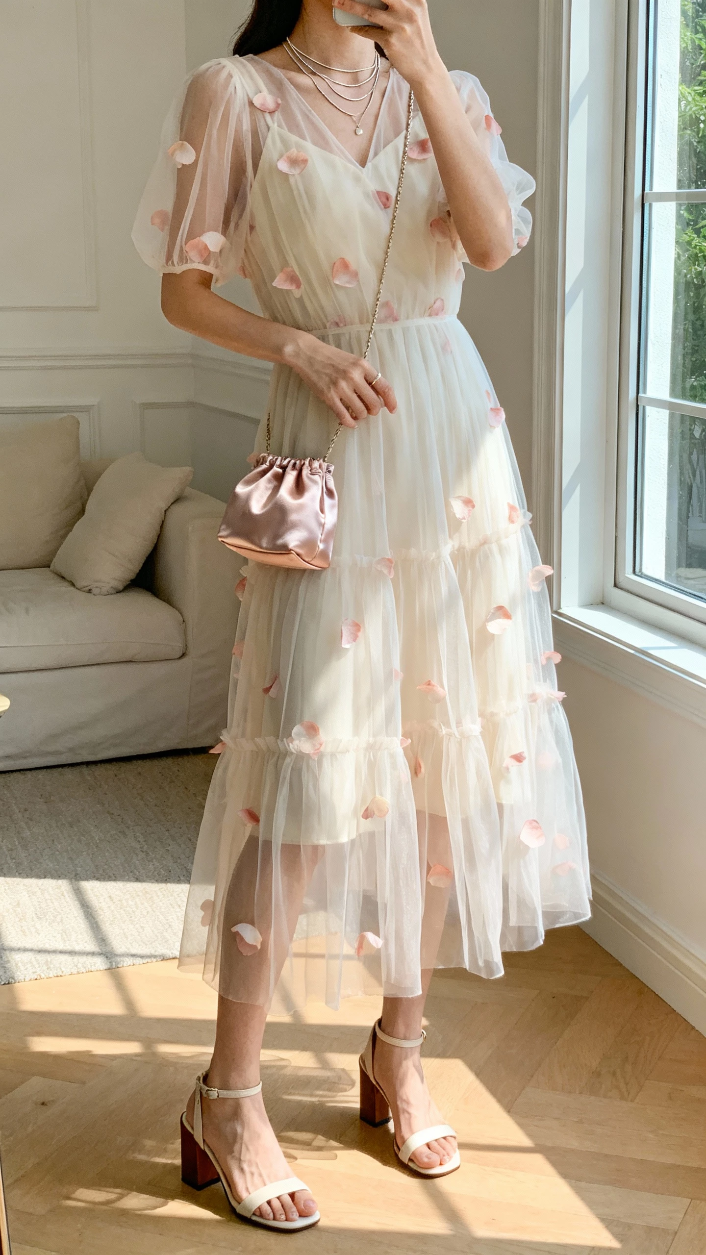 iPhone selfie cropped at neck showing a woman wearing an organza midi dress with sheer layers and petal appliqués, block-heel sandals, thin layered necklaces, and a satin mini bag, face not visible, airy living room by a window, bright natural light, casual iPhone aesthetic.