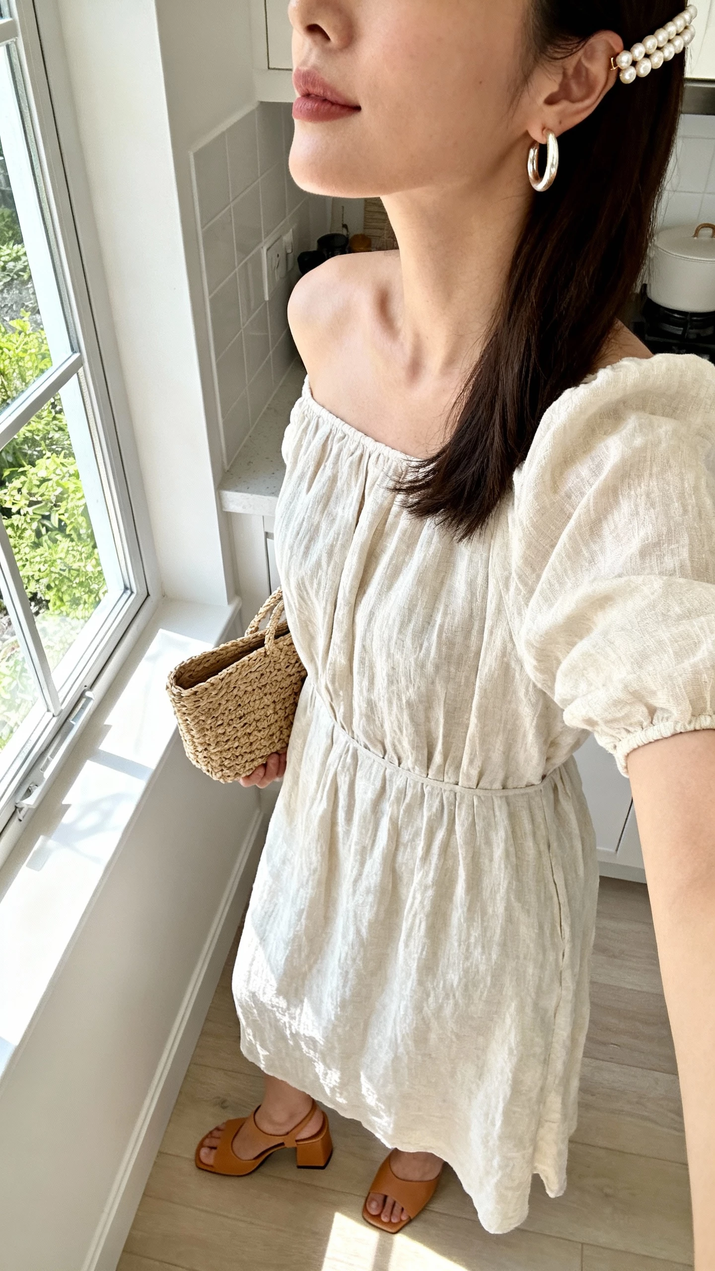 iPhone selfie cropped at shoulders of a woman in a breezy linen-blend midi dress, pearl-pop accents (pearl hoops and a pearl-accent hair clip), woven clutch, tan block-heel sandals, in a bright kitchen by a window, face not visible, soft morning light, natural iPhone quality.