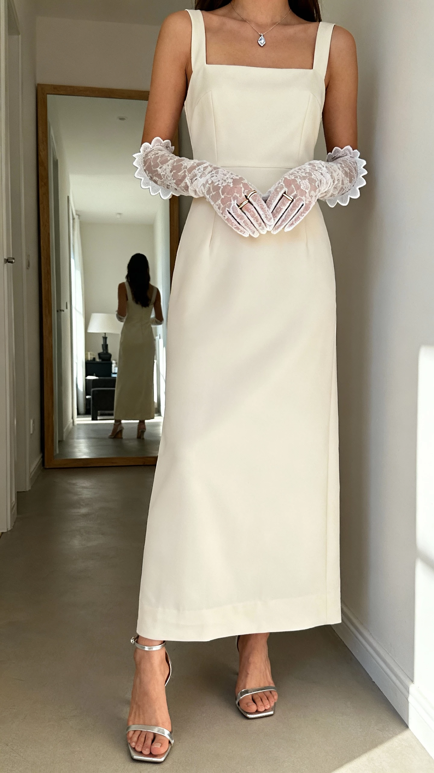 iPhone selfie cropped at shoulders of a woman wearing a clean crepe column gown, wrist-length Chantilly lace gloves with scalloped edges, delicate ankle-strap heels in metallic, a fine diamond pendant, and slim stacking rings; face not visible, hallway mirror in a minimalist apartment, morning light, natural iPhone photo quality.