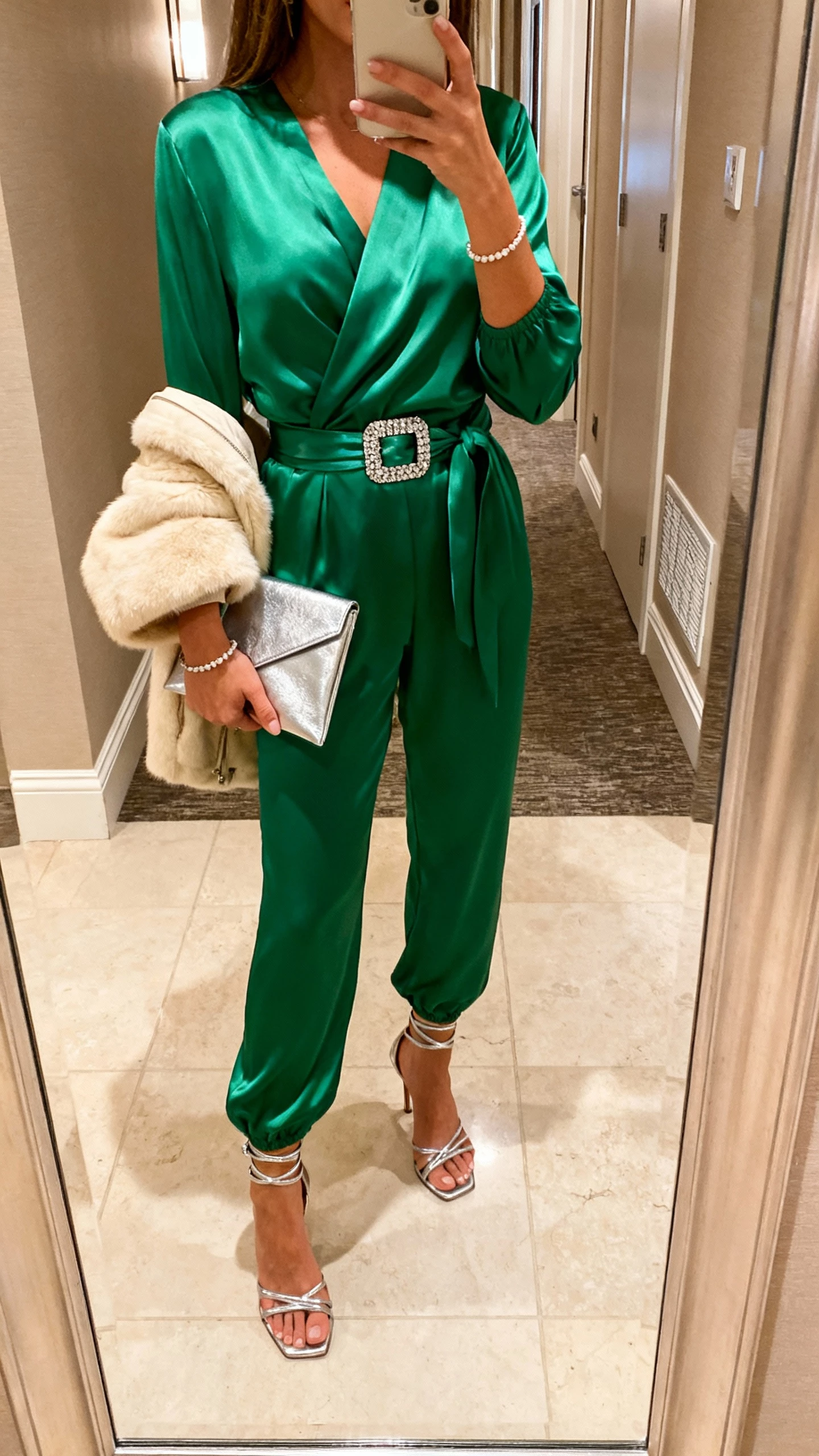 iPhone selfie cropped at shoulders showing a woman in an emerald satin wrap jumpsuit with long sleeves, rhinestone buckle belt, silver strappy heeled sandals, dainty tennis bracelet, silver envelope clutch, cream faux-fur cropped jacket over forearm; face not visible, hotel hallway mirror, warm indoor lighting, natural iPhone aesthetic.