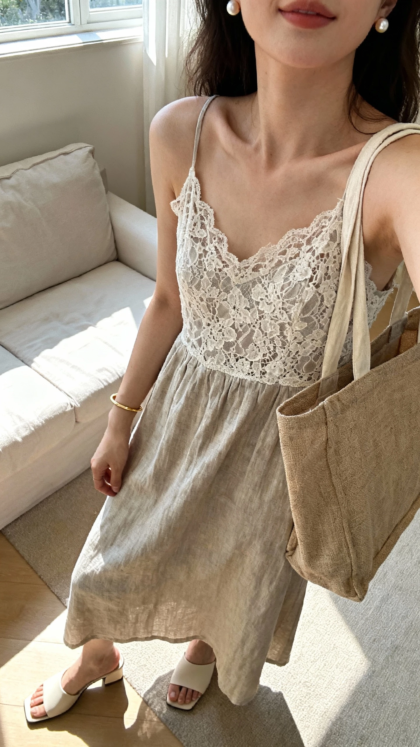 iPhone selfie cropped at shoulders showing a woman in a lace-meets-linen midi dress, minimalist heeled mules, pearl studs, thin gold bracelet, linen tote, face not visible, airy living room near a window with morning light, natural iPhone aesthetic.