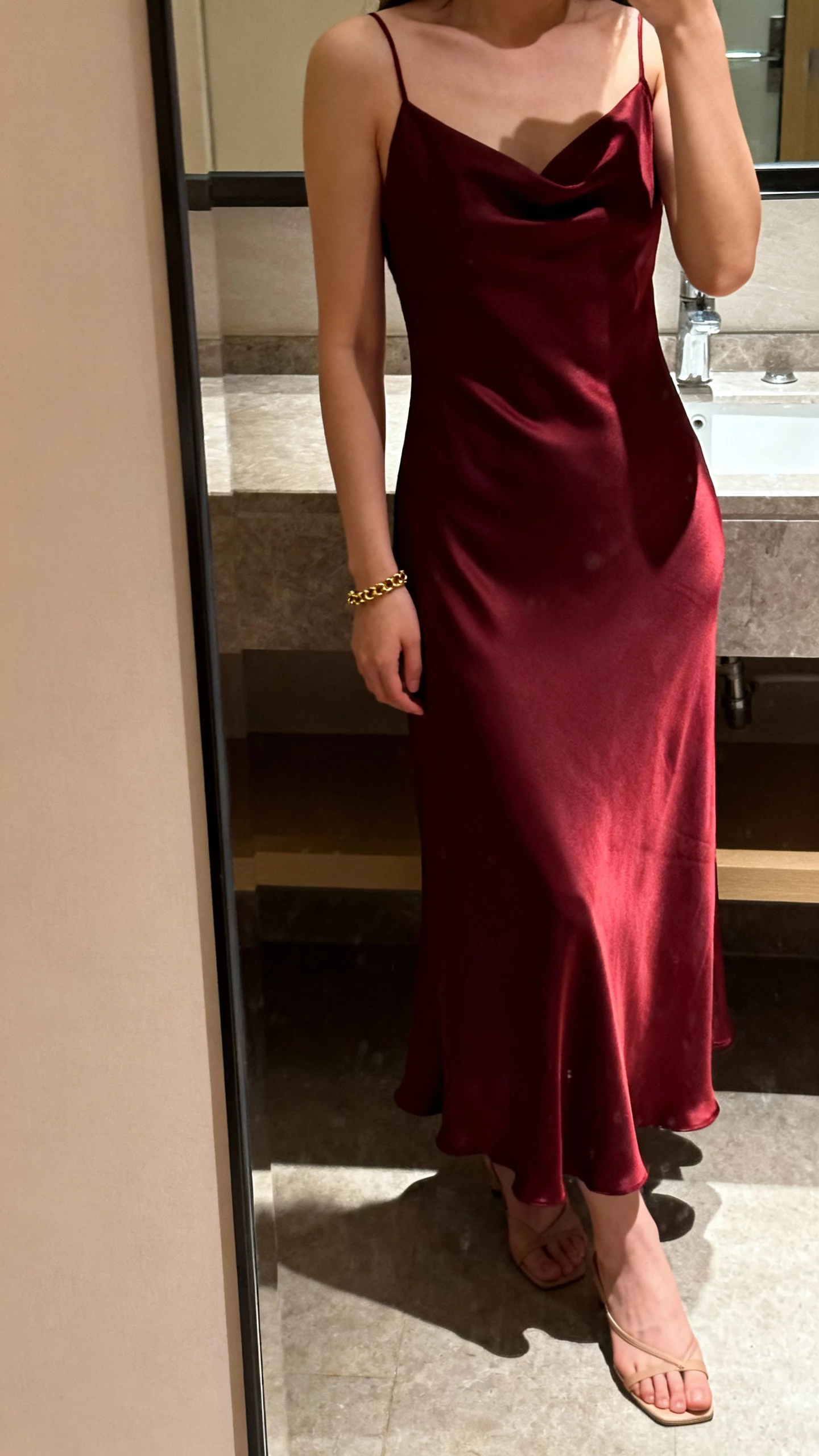 iPhone selfie cropped at shoulders showing a woman in a burgundy satin slip dress with liquid shine, delicate gold chain bracelet, minimalist heels, face not visible, bathroom mirror, evening indoor lighting, natural iPhone aesthetic.