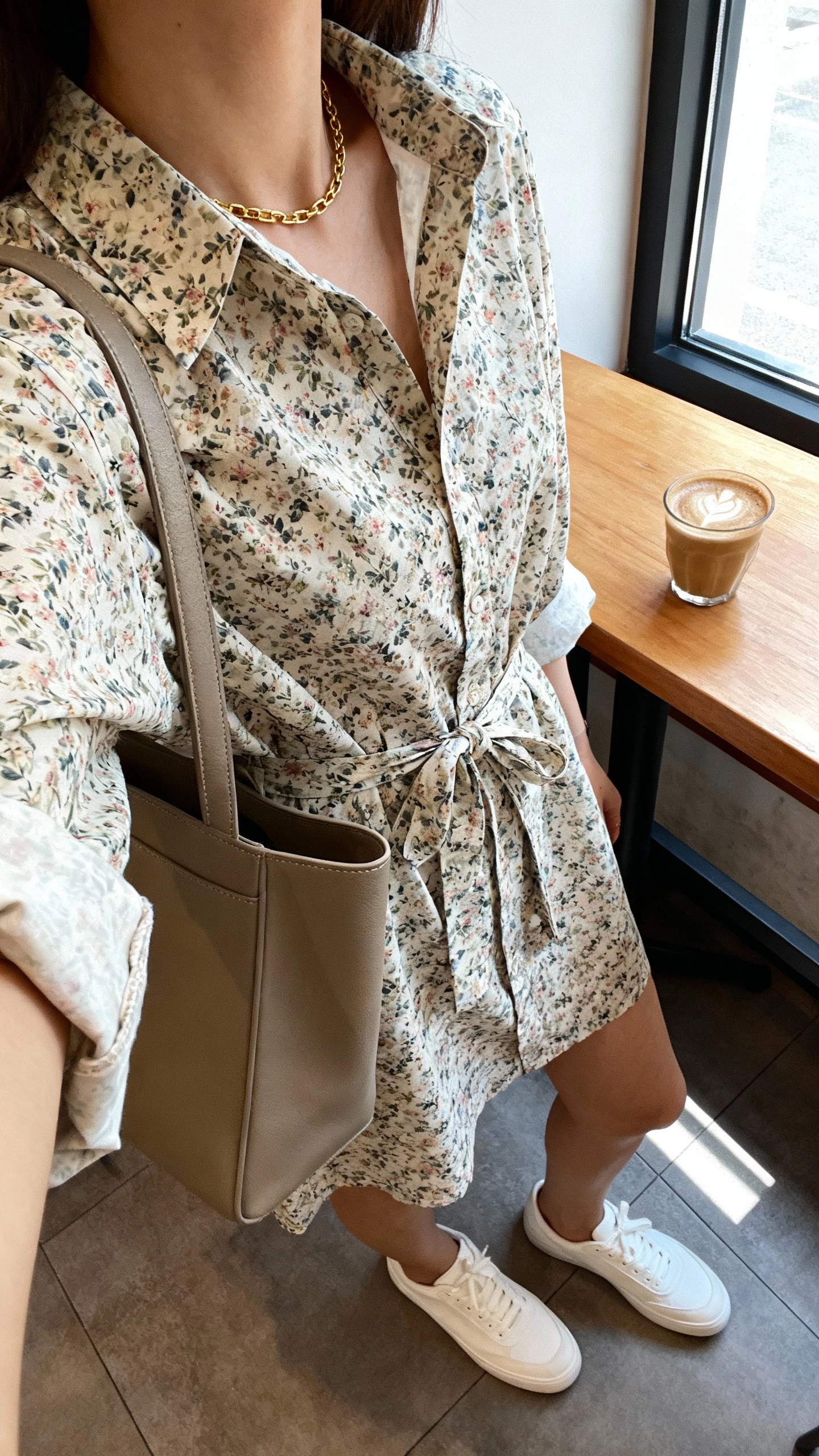 iPhone selfie cropped at shoulders showing a woman in a floral shirt-dress with a tie waist, minimalist white low-top sneakers, thin gold chain necklace, and a structured tote, face not visible, coffee shop table shot, morning window light, natural iPhone quality.