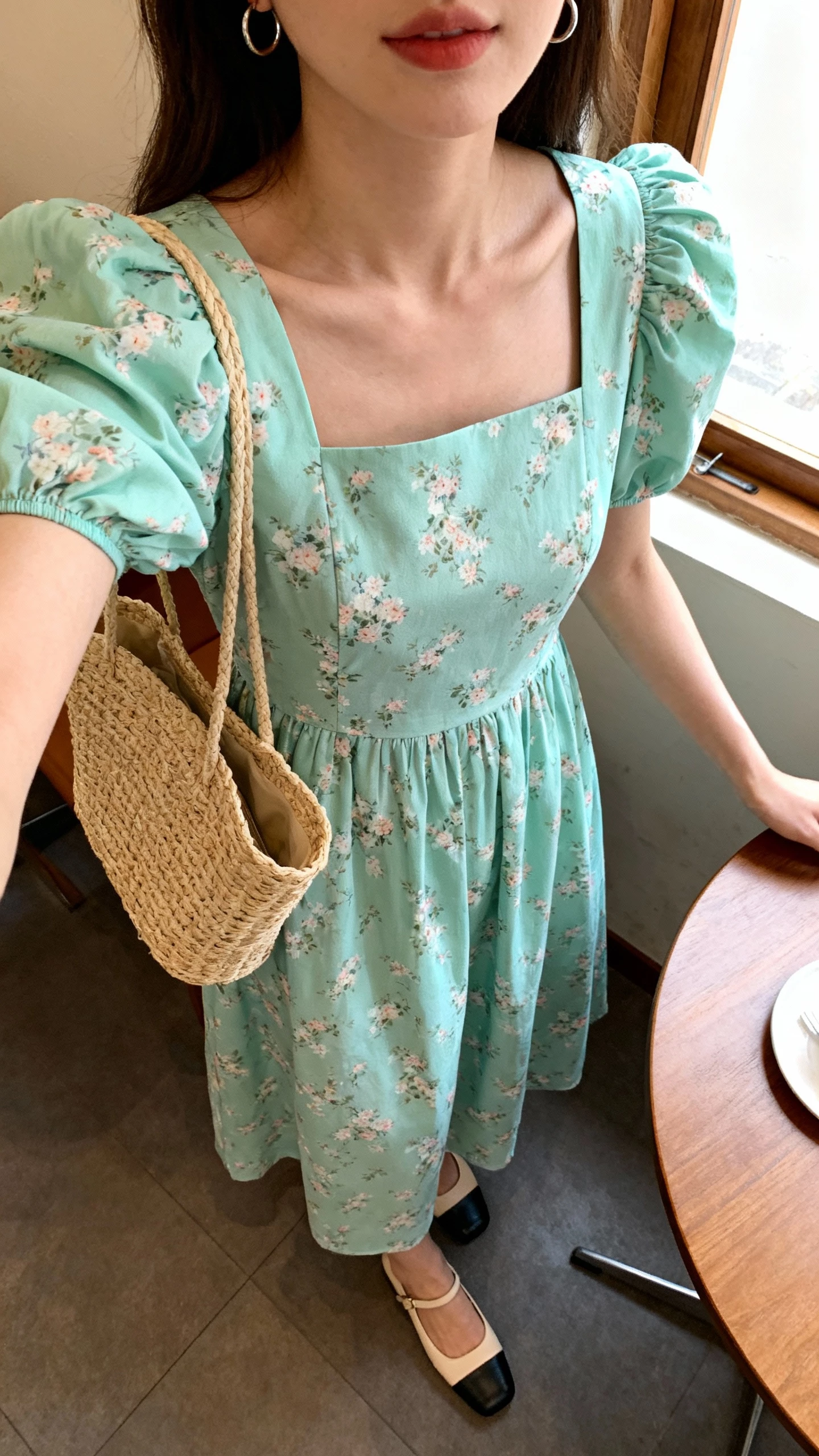 iPhone selfie cropped at shoulders showing a woman in a mint floral puff-sleeve tea dress with square neckline, low block heels, straw bag, small hoops, face not visible, cafe table by a window with soft daylight, natural iPhone photo quality.