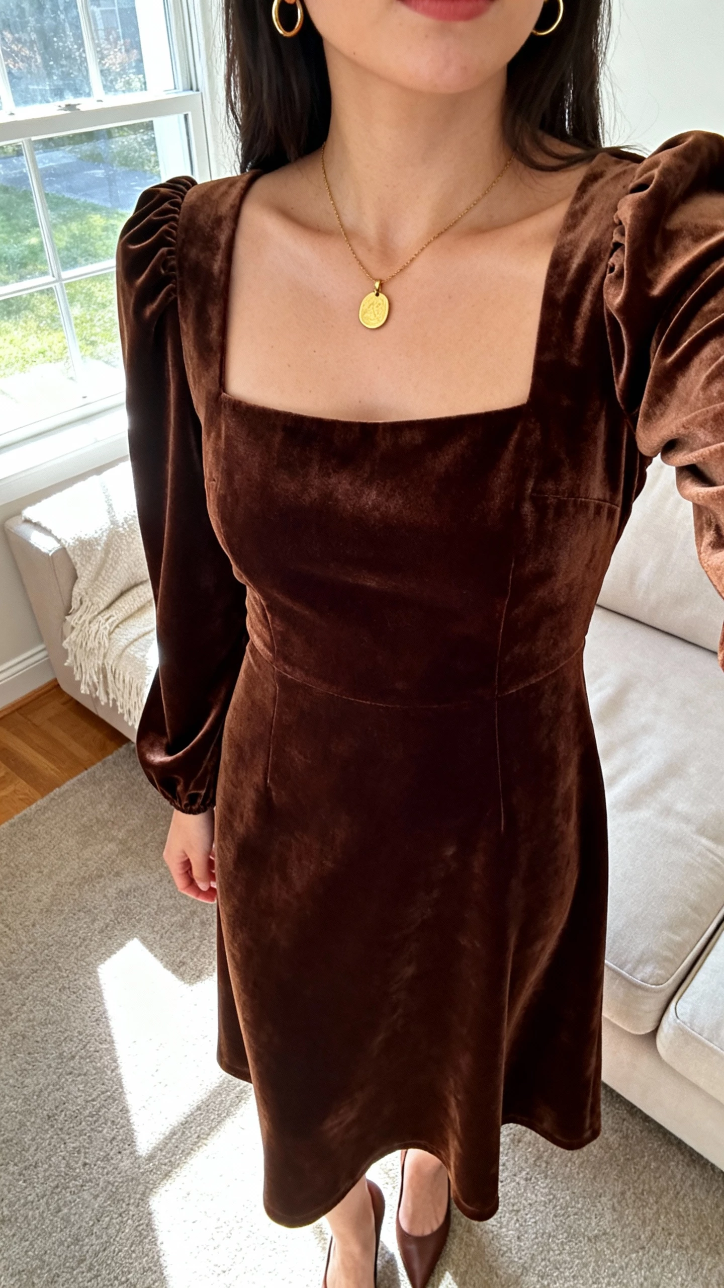 iPhone selfie cropped at shoulders showing a woman in a chocolate brown velvet dress with a square neck and subtle puff sleeves, gold pendant and small hoops, and classic heels, face not visible, cozy living room, morning window light, natural iPhone aesthetic.