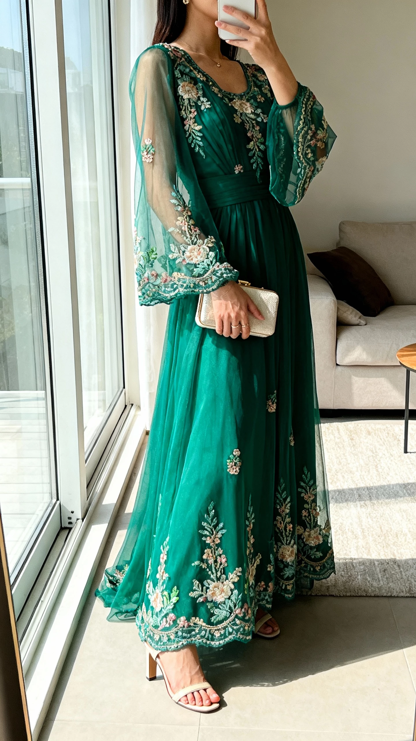 iPhone side-angle selfie cropped at shoulders showing a woman wearing an emerald embroidered gown with sheer sleeves, small clutch, heeled sandals, face not visible, living room by window, natural daylight, iPhone photo quality.