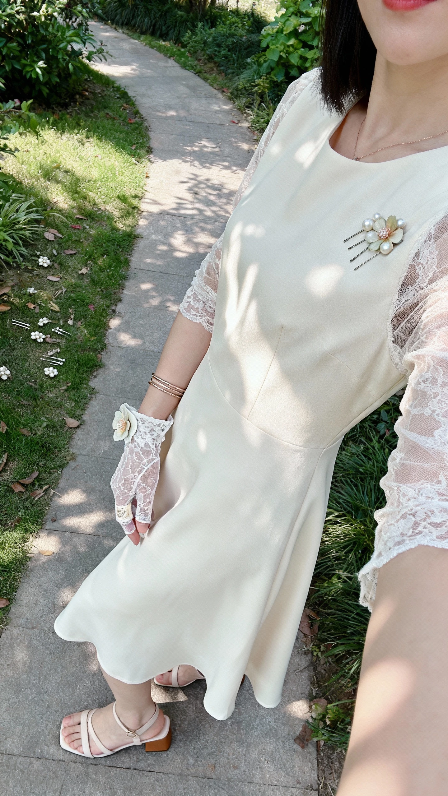 iPhone side-angle selfie of a woman in a flowing A-line dress with sheer lace sleeves, fingerless lace gloves with a thumb loop, low block-heel sandals, scattered hair pins with pearl/pressed-flower detail, and fine stacked bracelets; face not visible, garden path setting, dappled afternoon light, casual iPhone photo.