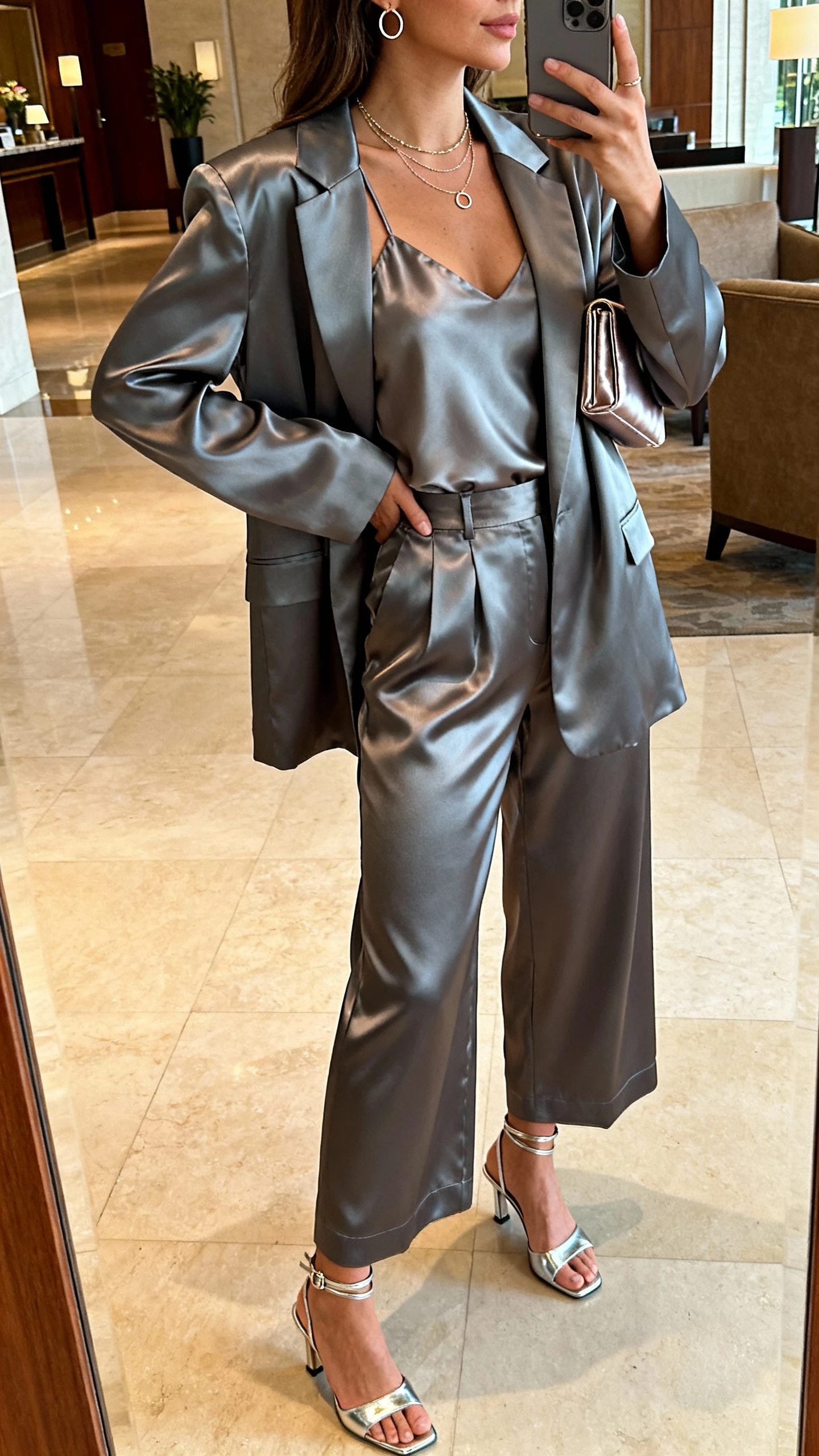 iPhone side-angle selfie of a woman in a gunmetal relaxed-fit satin blazer, matching wide-leg high-waist satin trousers, strappy satin camisole, metallic ankle-strap heels, layered delicate necklaces with dainty hoops, slim satin clutch; hotel lobby mirror, face not visible, warm indoor lighting, natural iPhone photo quality.