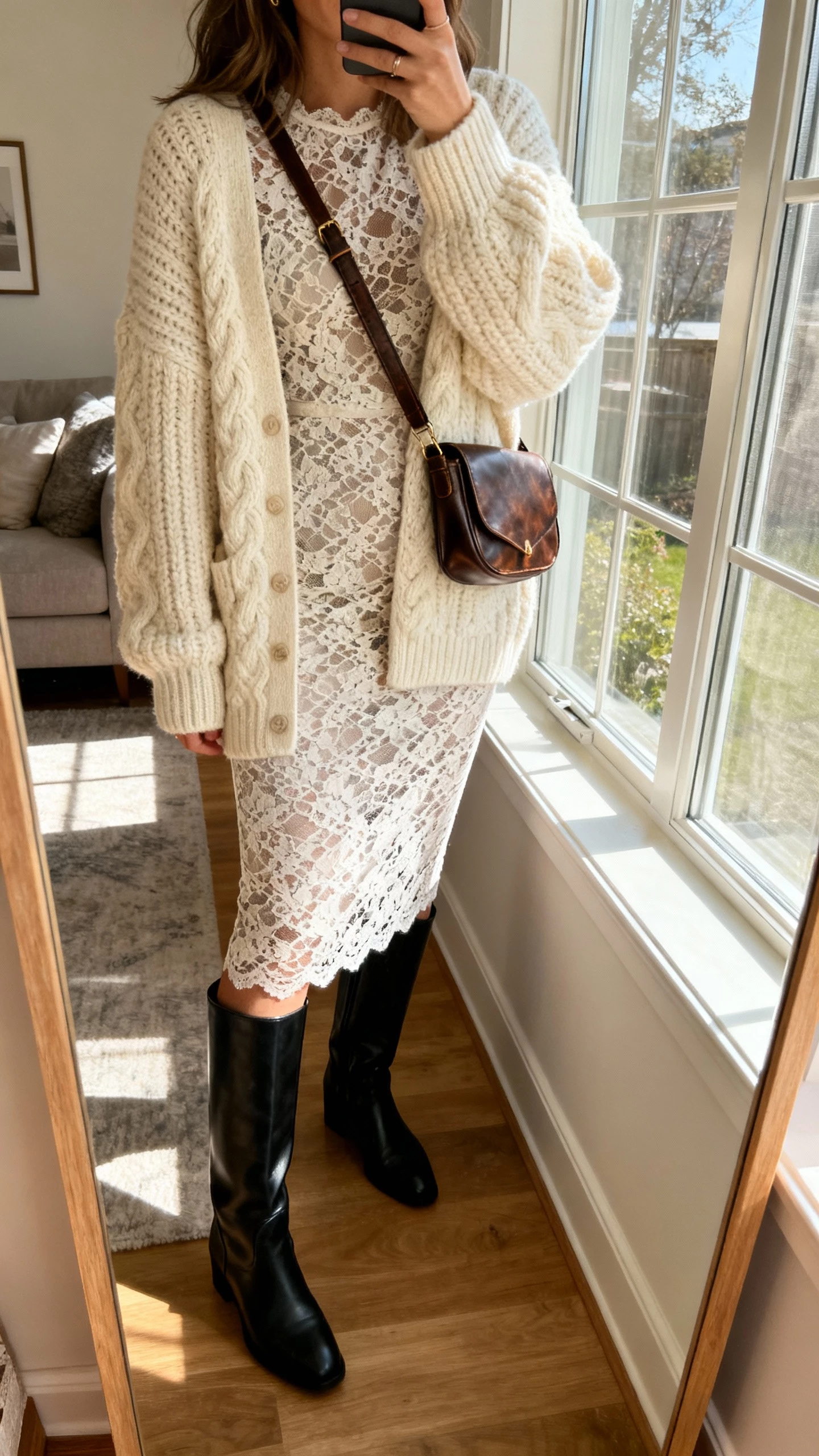 iPhone side-angle selfie of a woman in a lace pencil dress layered with a chunky cream knit cardigan, black knee-high boots, and a leather crossbody; face not visible, cozy living room mirror, afternoon window light, natural iPhone photo.