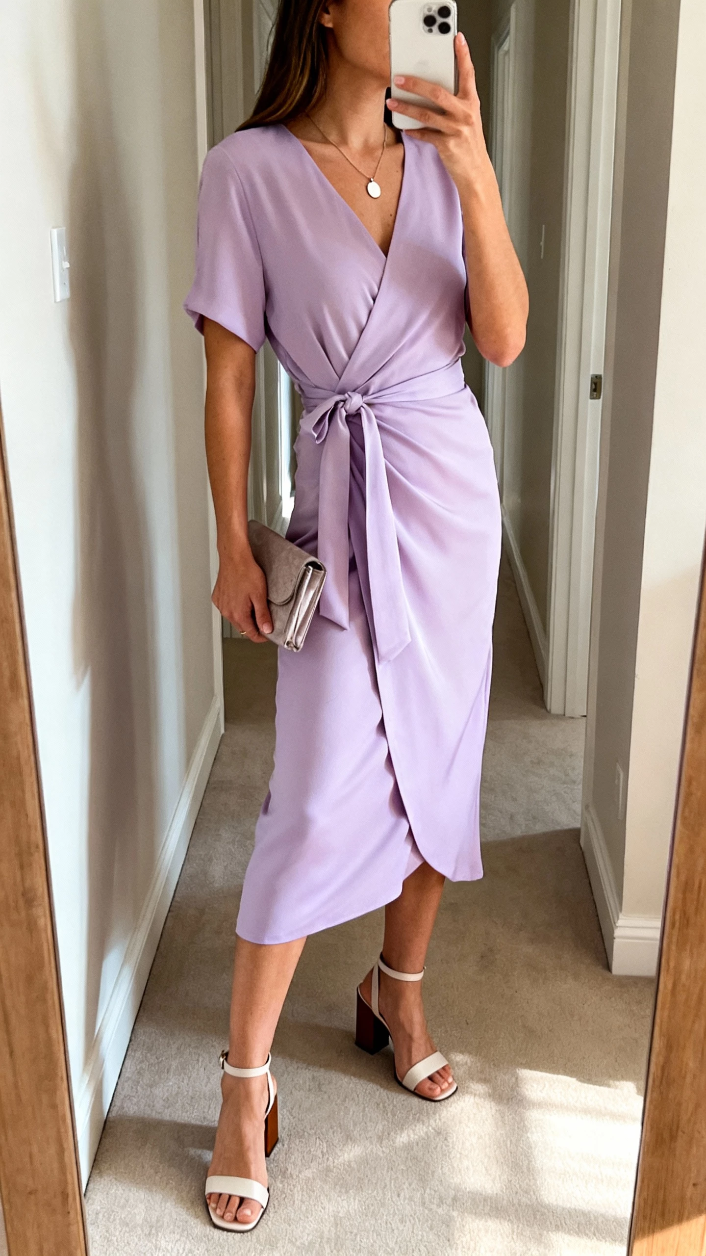 iPhone side-angle selfie of a woman in a lavender wrap midi dress with tie waist, block-heel sandals, simple pendant necklace, small clutch, face not visible, taken in a hallway mirror with afternoon light, casual iPhone photo.