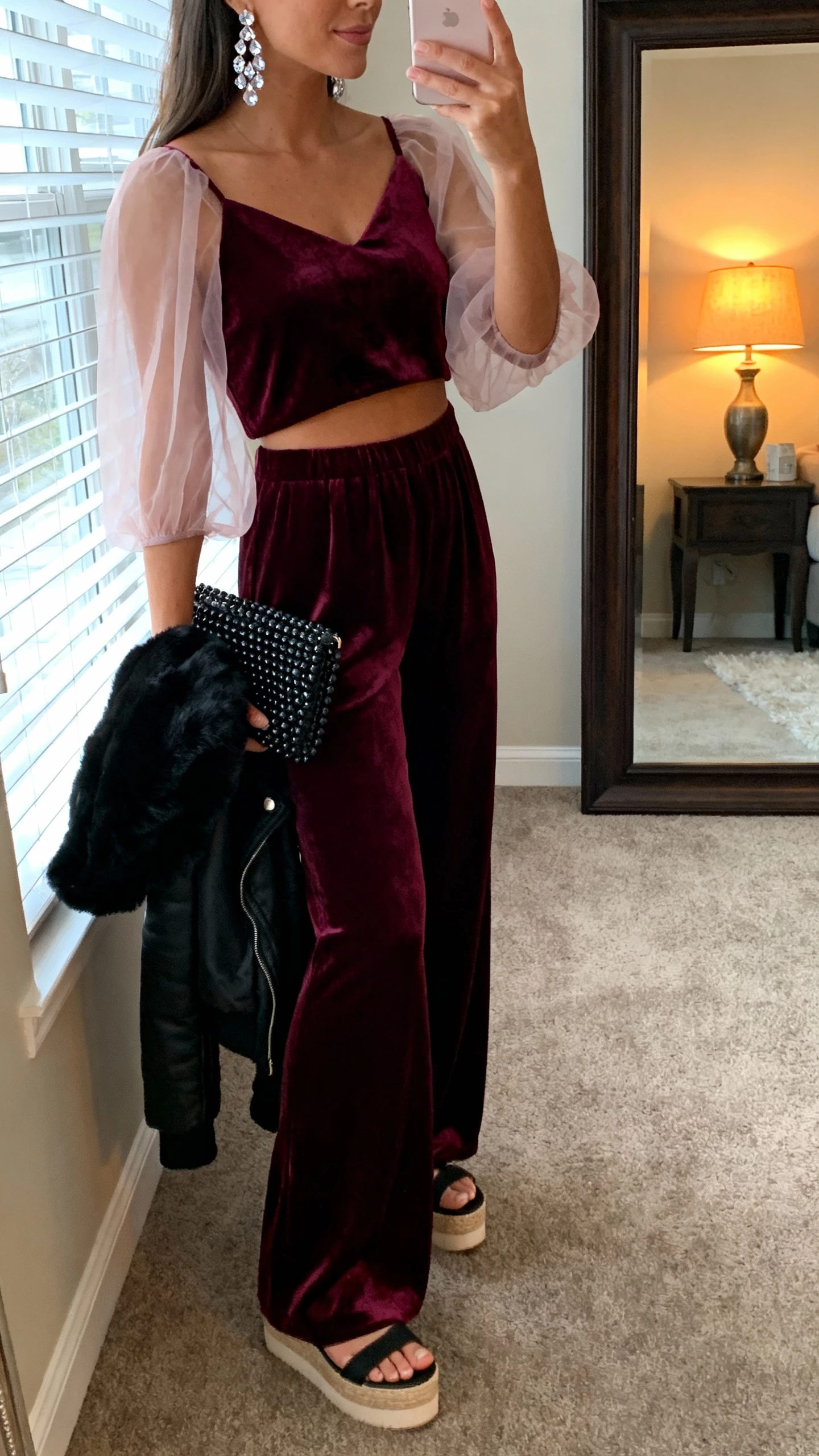 iPhone side-angle selfie of a woman in a merlot velvet palazzo jumpsuit with sheer organza sleeves, crystal drop earrings, platform sandals, black beaded clutch, cropped black faux-fur jacket draped over forearm; face not visible, living room mirror, warm lamplight and window glow, natural iPhone aesthetic.