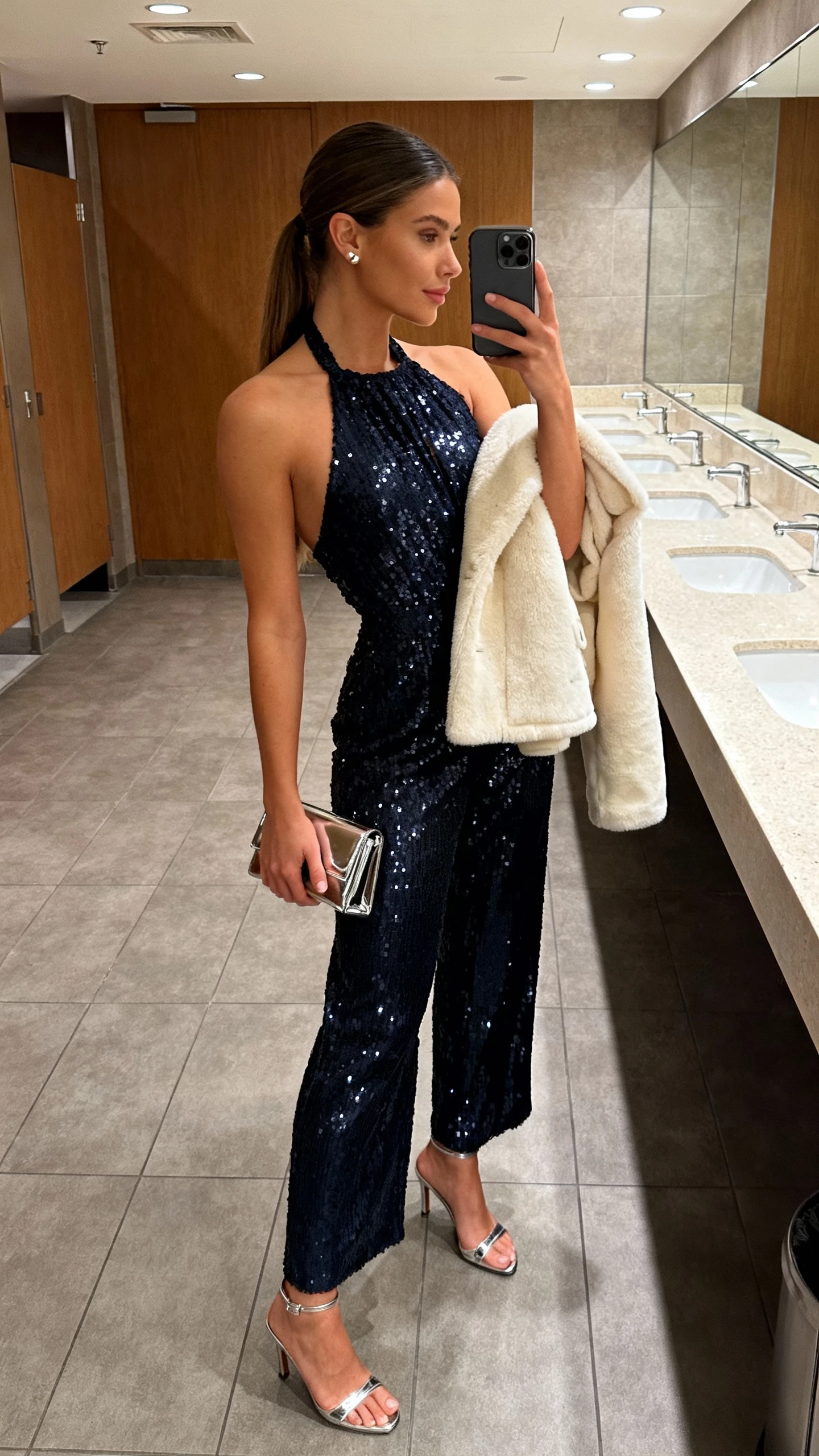 iPhone side-angle selfie of a woman in a midnight sequin halter jumpsuit, minimalist metallic ankle-strap heels, delicate stud earrings, glossy micro clutch, cream faux shearling short coat tossed over shoulder; face not visible, bathroom mirror at venue, warm overhead lighting, natural iPhone quality.