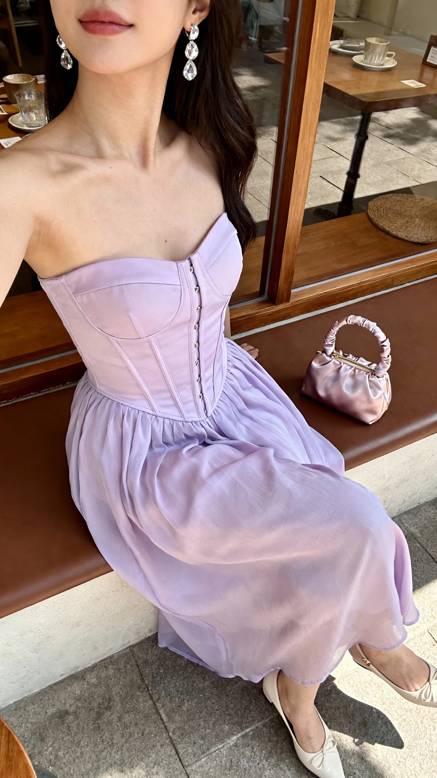 iPhone side-angle selfie of a woman in a powder-lilac set with a corset top and flowing skirt, kitten heels, crystal earrings, and a satin mini bag, face not visible, captured by a cafe window seat, morning light, natural iPhone photo quality.