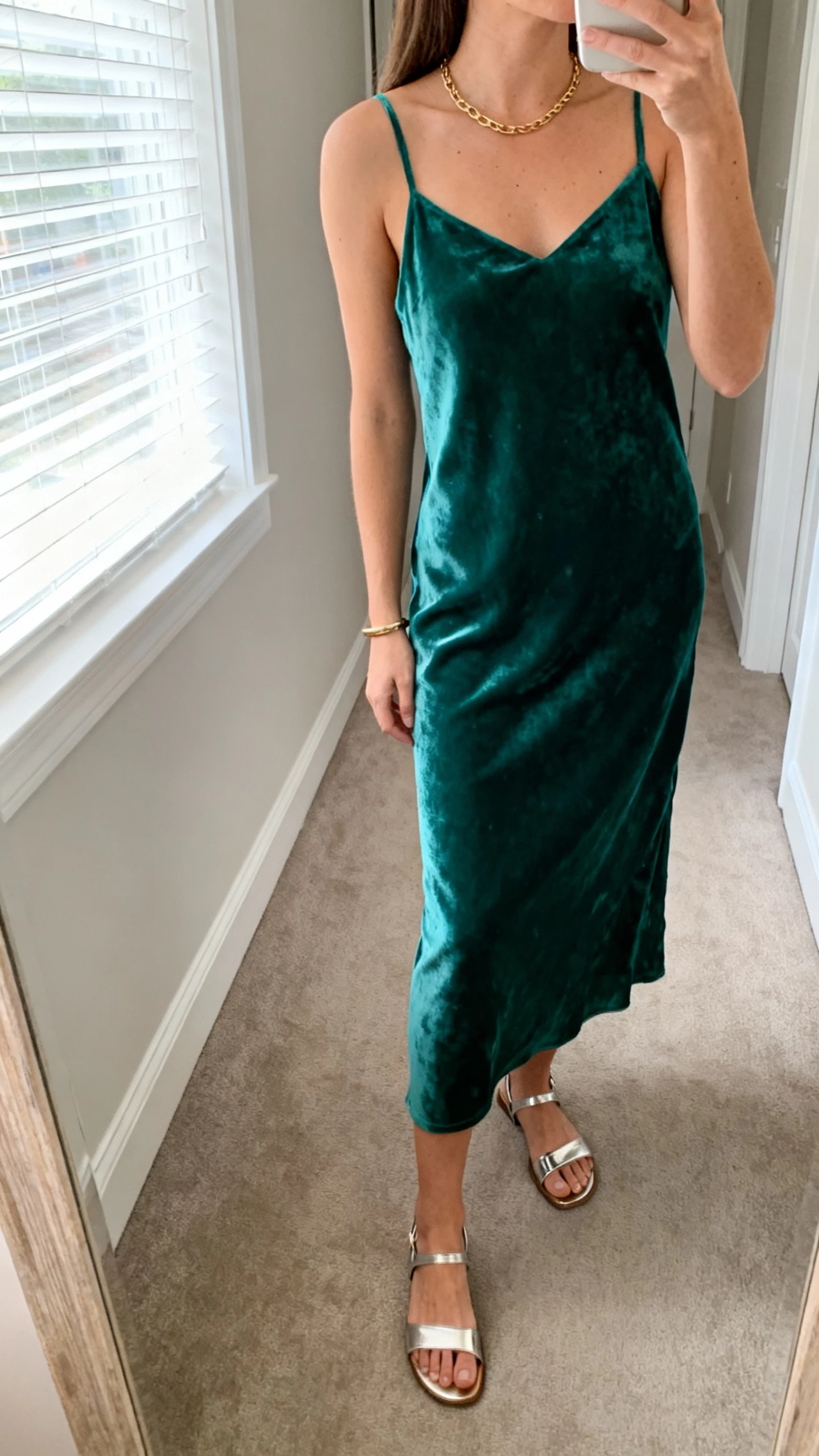 iPhone side-angle selfie of a woman in an emerald velvet slip midi that glides, minimal gold necklace, slim bracelet, metallic sandals, face not visible, hallway mirror, morning window light, natural iPhone aesthetic.