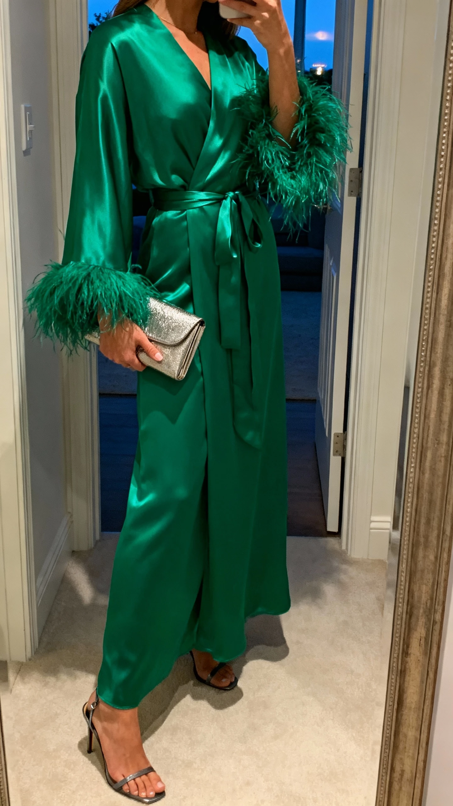 iPhone side-angle selfie of a woman in an emerald satin wrap gown with a feather-trim moment at the cuffs, metallic clutch, slingback heels, face not visible, entryway mirror, evening indoor light, natural iPhone photo.