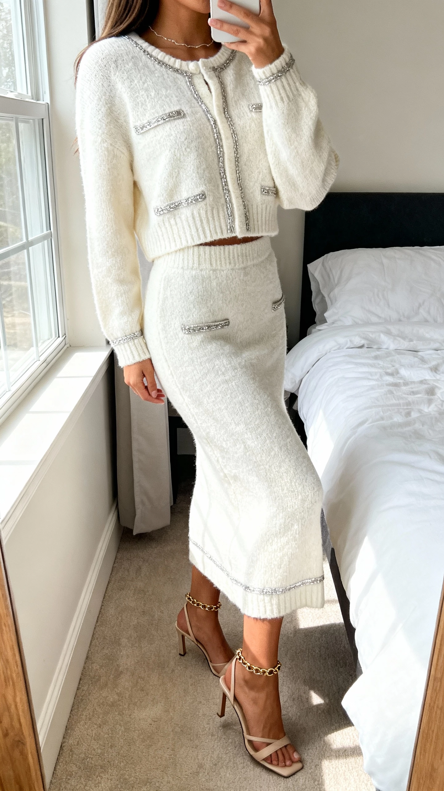 iPhone side-angle selfie of a woman in an ivory knit skirt set with soft metallic accents, delicate chain, slingback heels, face not visible, bedroom near window, gentle natural light, casual iPhone photo