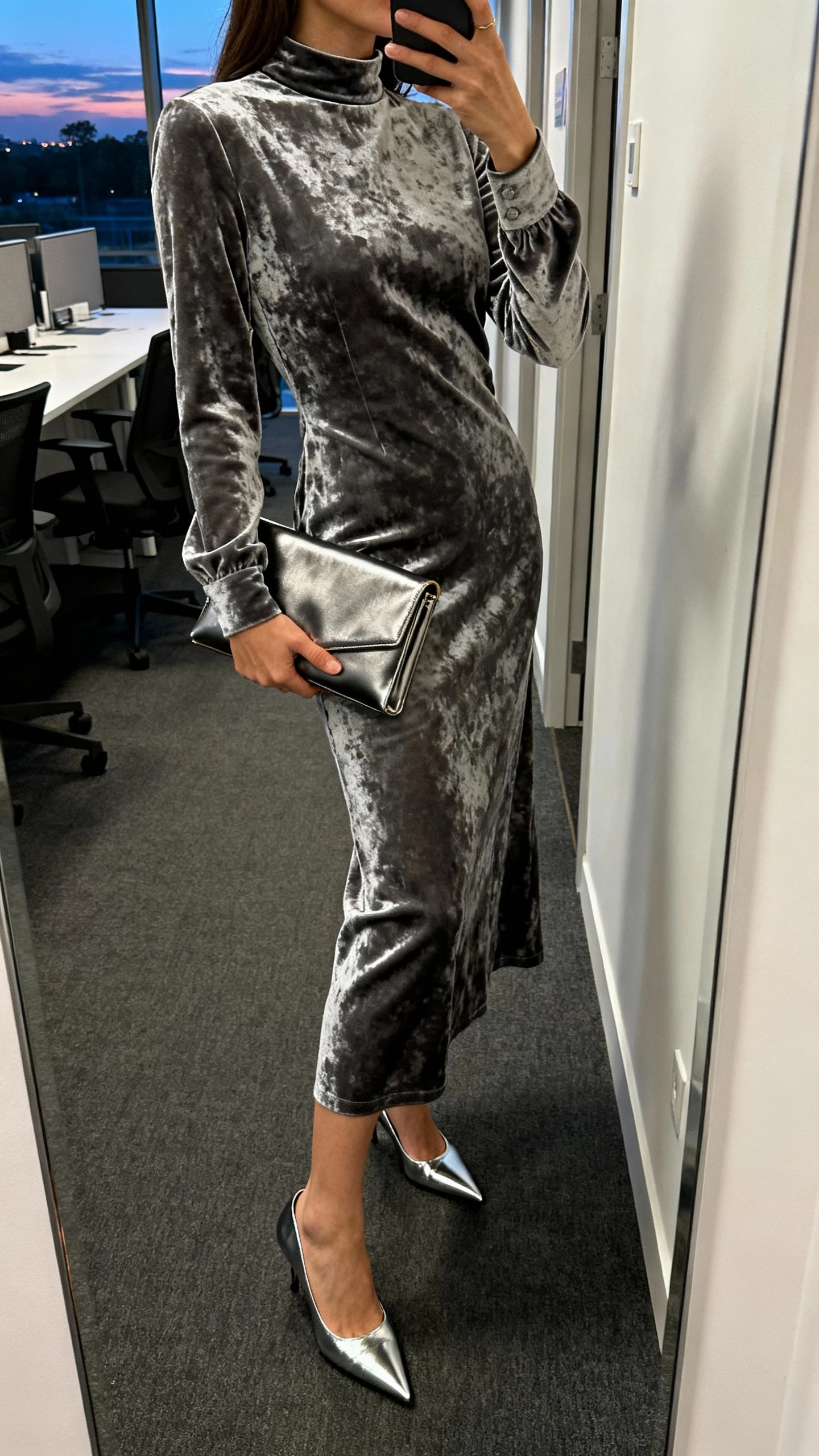 iPhone side-angle selfie of a woman in gunmetal crushed velvet dress with a mock neck and long sleeves, sleek clutch, and pointed metallic heels, face not visible, office corridor mirror at dusk, natural indoor lighting, iPhone photo quality.