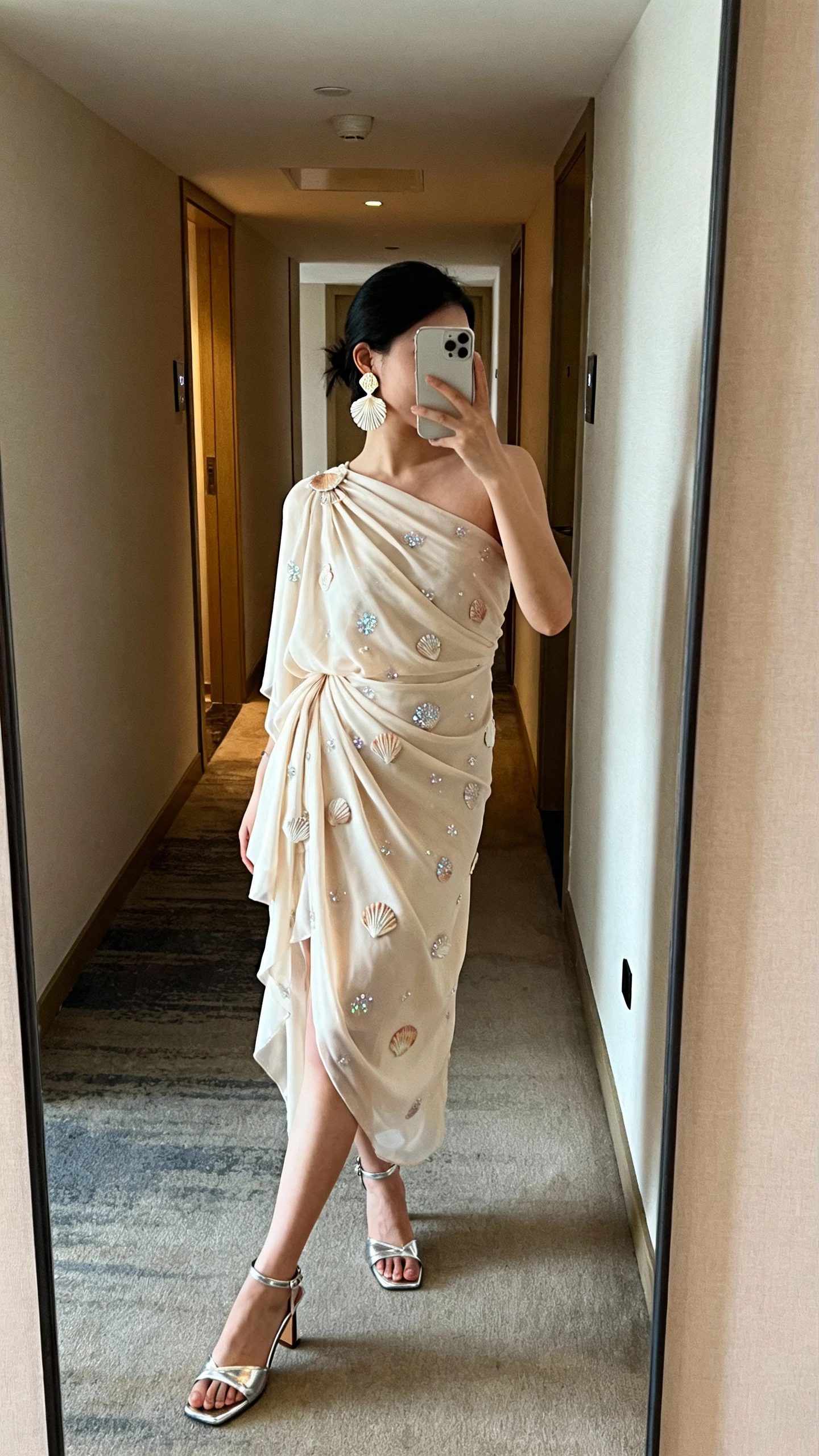 iPhone side-angle selfie of a woman wearing a one-shoulder chiffon dress with seashell sparkle details, metallic heeled sandals, shell-inspired earrings, face not visible, hotel hallway mirror, soft indoor lighting, natural iPhone aesthetic.