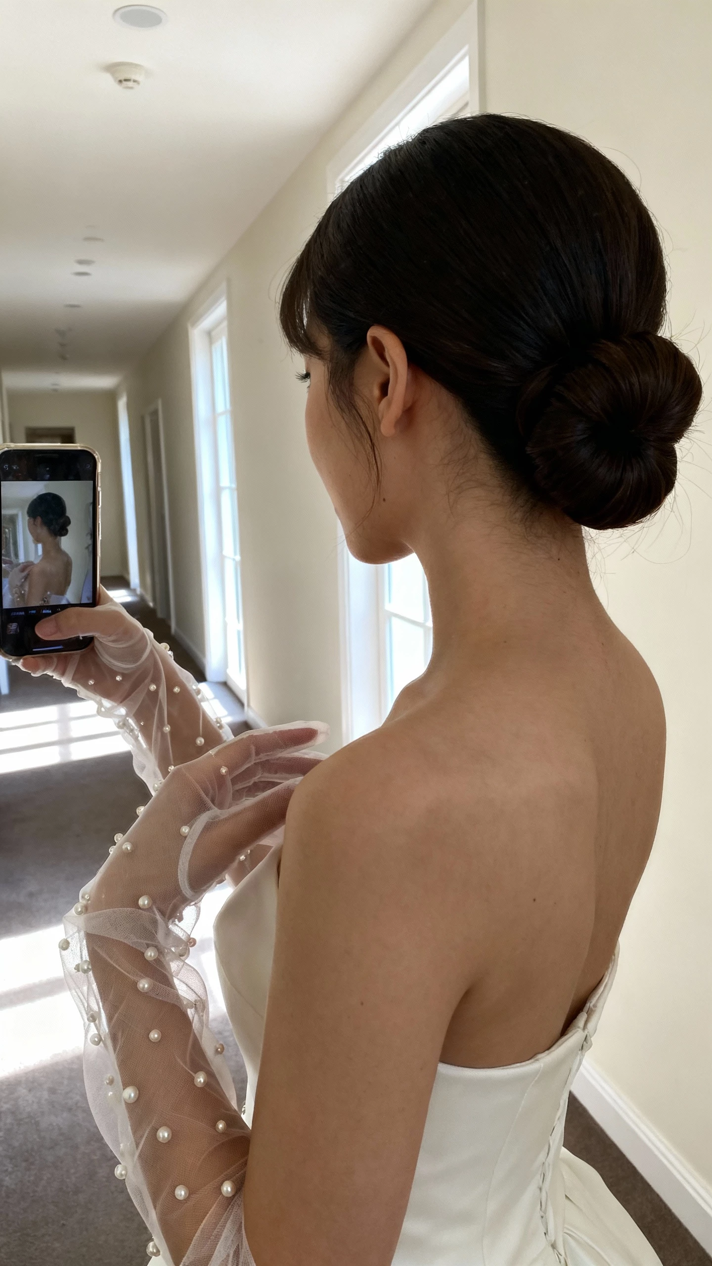 iPhone side-angle selfie of a woman wearing sheer tulle gloves with tiny pearls and a classic chignon, paired with a simple bridal dress, face not visible, hallway mirror, morning natural light, casual iPhone photo