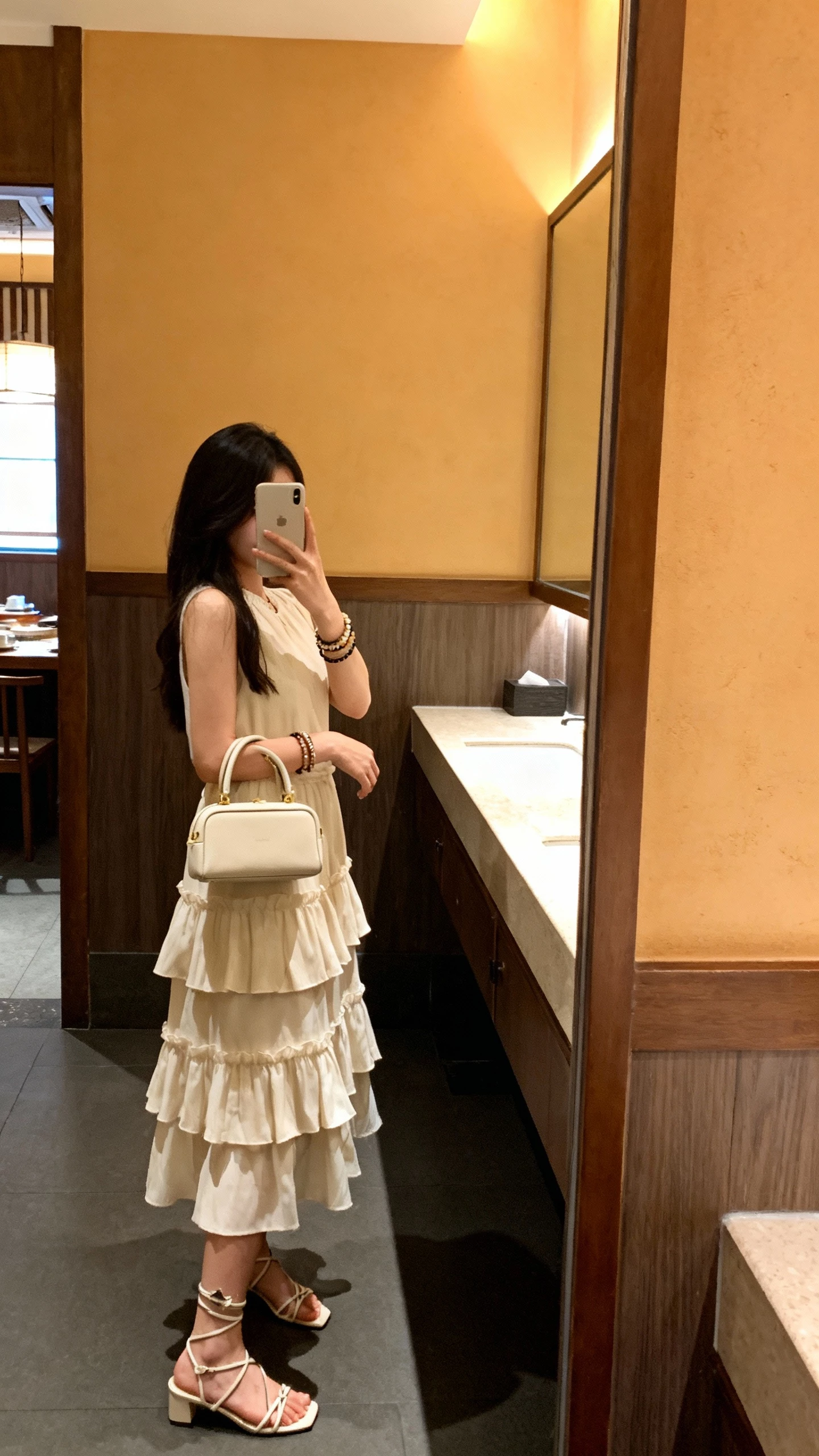 iPhone side mirror selfie of a woman wearing a tiered ruffle midi dress, strappy heels, layered bracelets, and a petite top-handle bag, face not visible, bathroom mirror at a restaurant, warm indoor lighting, natural iPhone photo quality.