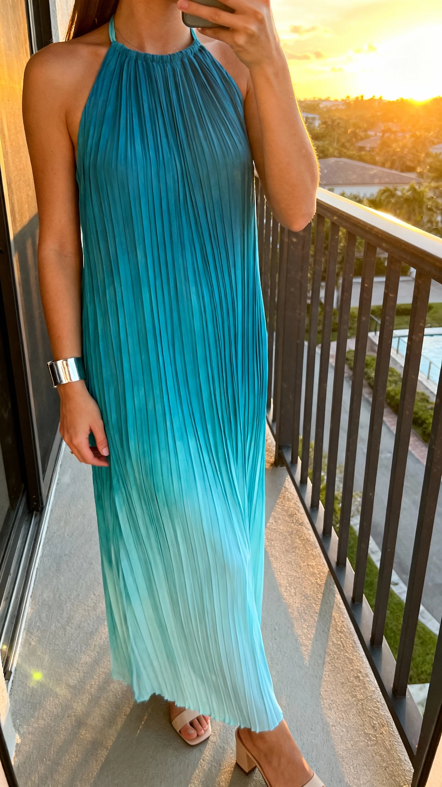iPhone side-profile selfie of a woman in a pleated halter dress with ocean-blue ombre, minimalist heels, silver cuff bracelet, face not visible, balcony at sunset, natural light, iPhone photo quality.