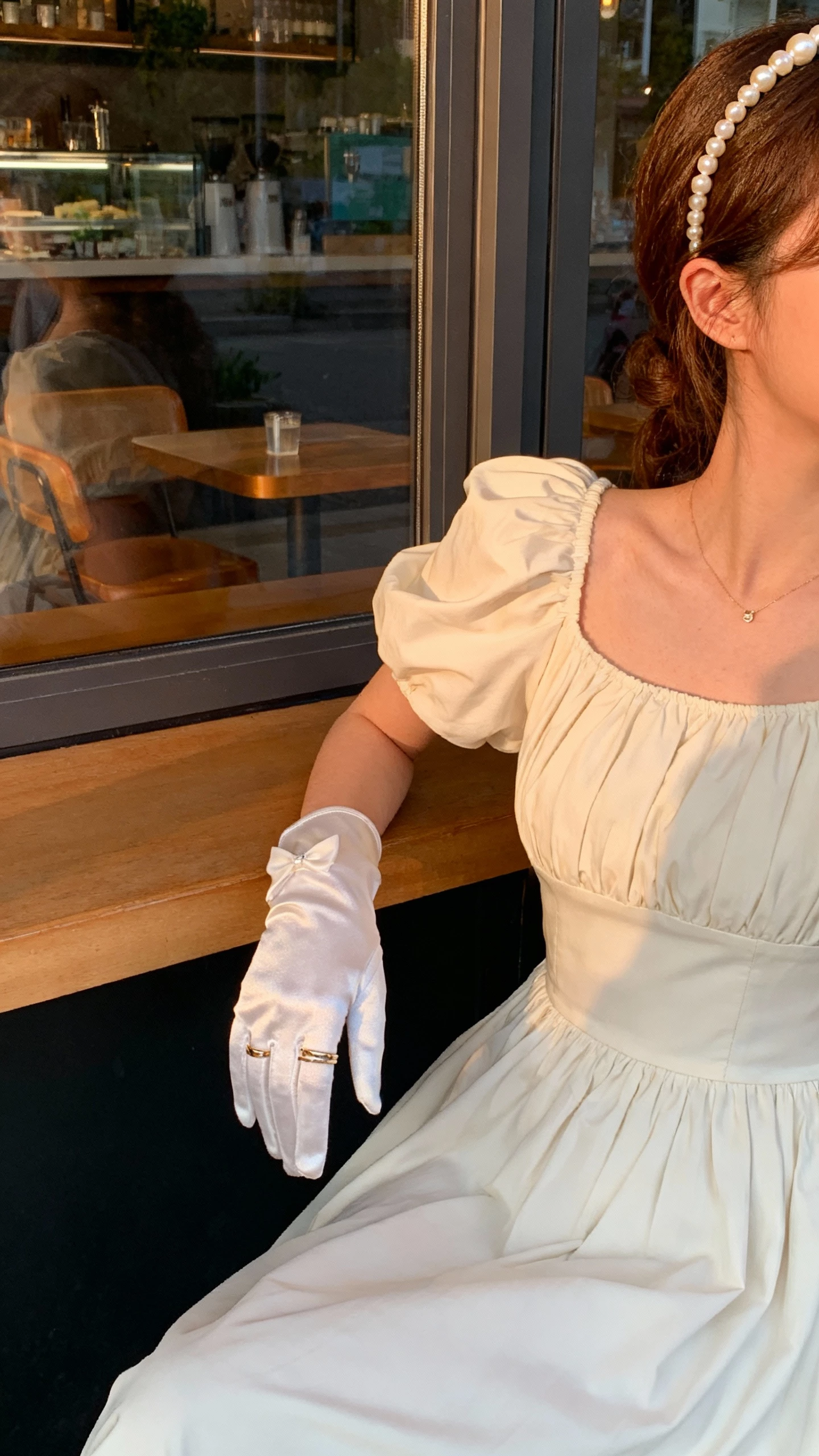 iPhone side-profile selfie of a woman wearing soft white opera gloves with a puff-sleeve empire-waist dress, pearl headband, and dainty rings, face not visible, taken near a cafe window seat, late afternoon natural light, natural iPhone aesthetic.