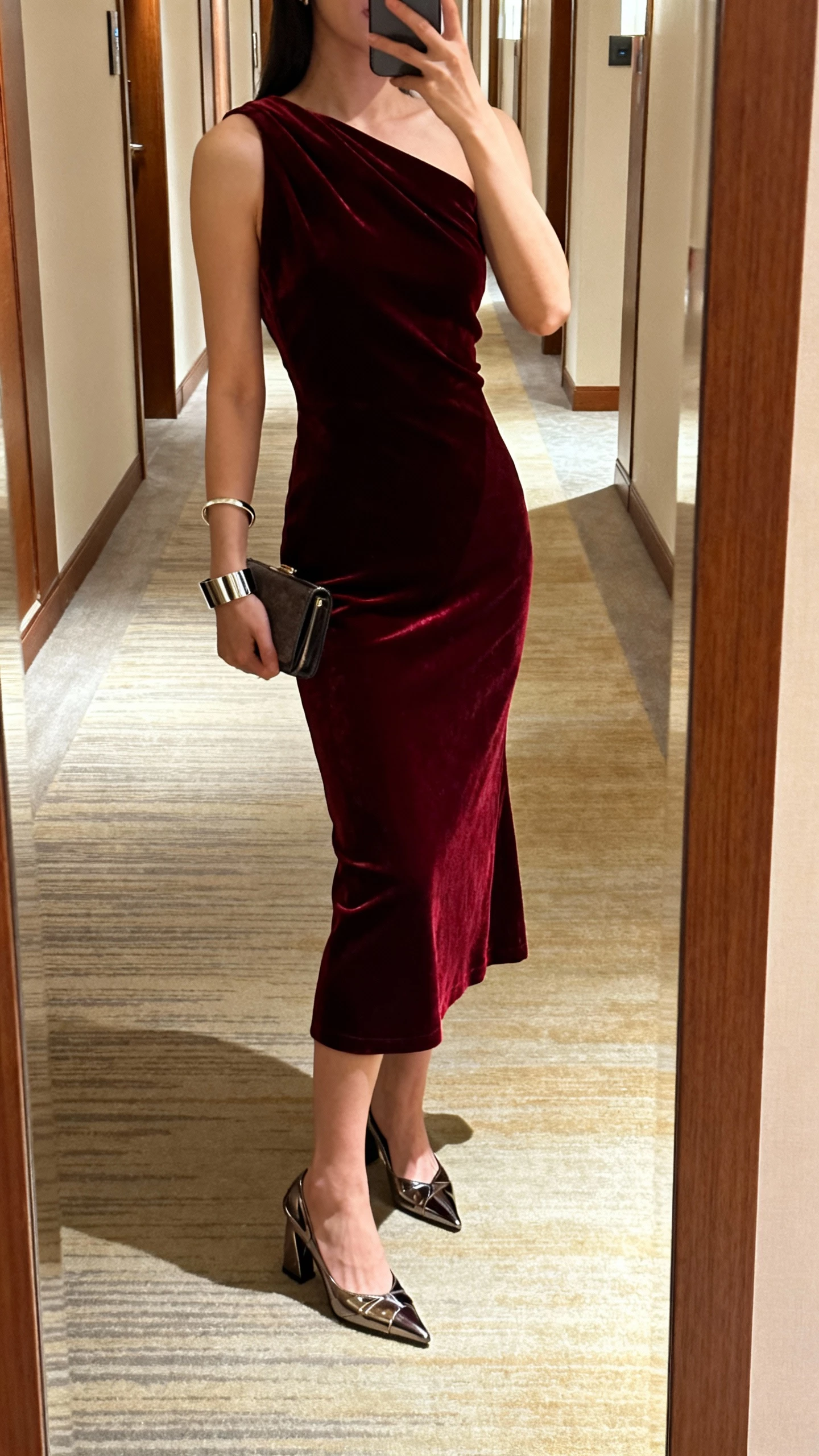 iPhone side-view selfie of a woman in a garnet velvet one-shoulder midi paired with sculptural metallic heels, sleek cuff bracelet, small clutch, face not visible, hotel hallway mirror, warm indoor lighting, casual iPhone photo.