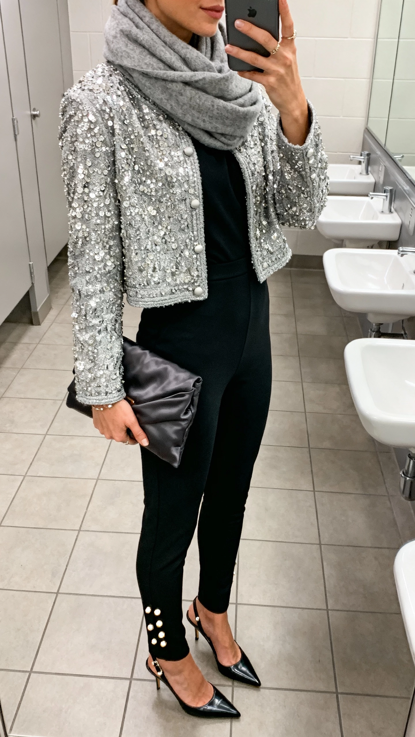 iPhone side-view selfie of a woman in a slim black ponte jumpsuit, silver beaded cropped jacket, pointed slingbacks, statement studs, charcoal satin clutch, soft gray wool-cashmere scarf looped at neck; face not visible, office restroom mirror, cool overhead lighting, natural iPhone quality.