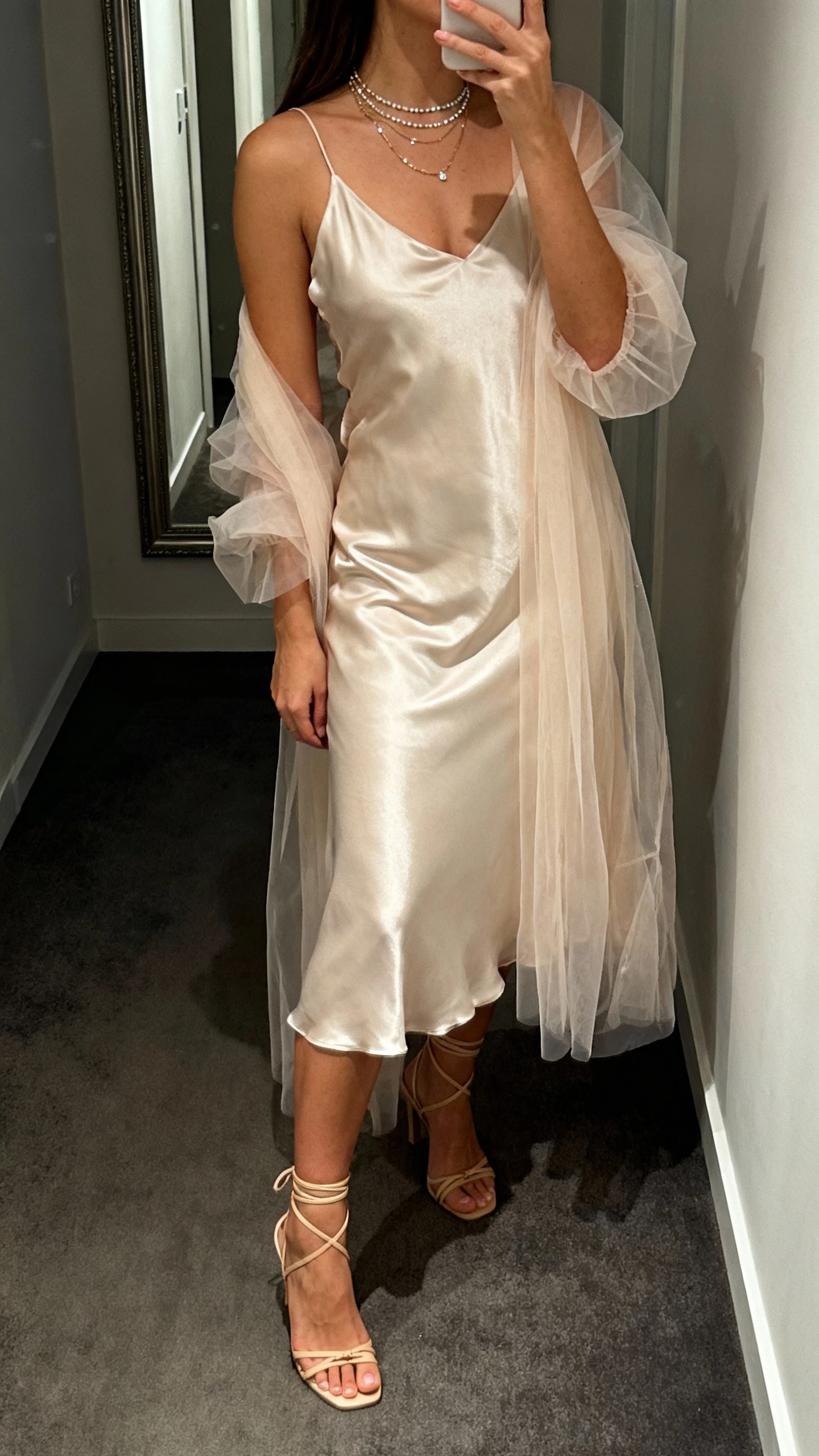 iPhone side-view selfie of a woman in a soft satin slip dress under a sheer tulle overlay, strappy heels, and delicate layered necklaces, face not visible, corridor mirror, evening indoor lighting, natural iPhone aesthetic