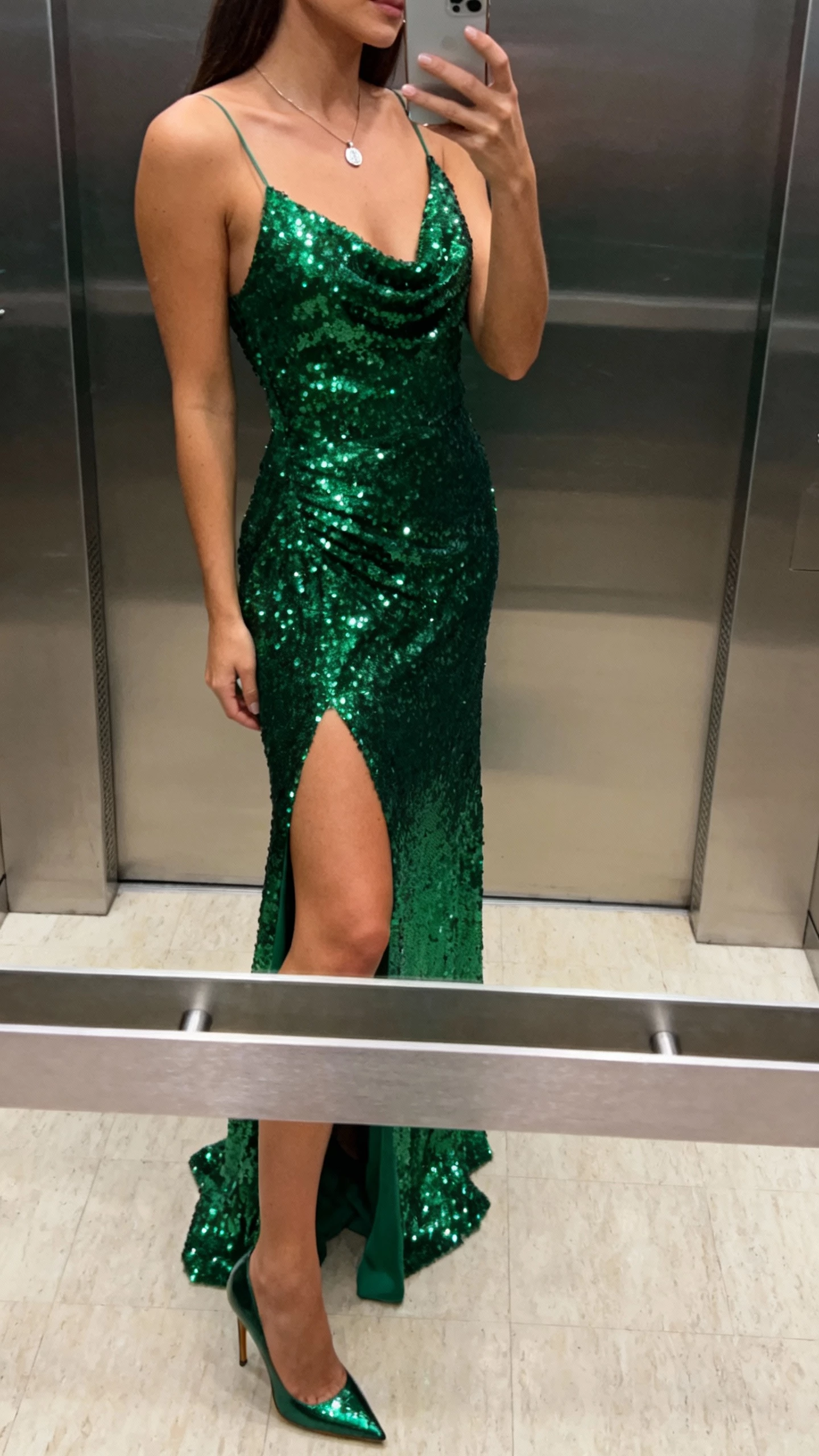 iPhone side-view selfie of a woman wearing an emerald sequined gown with a high slit for dance-floor glitter, simple pendant necklace, stiletto heels, face not visible, elevator mirror, indoor lighting, casual iPhone shot.
