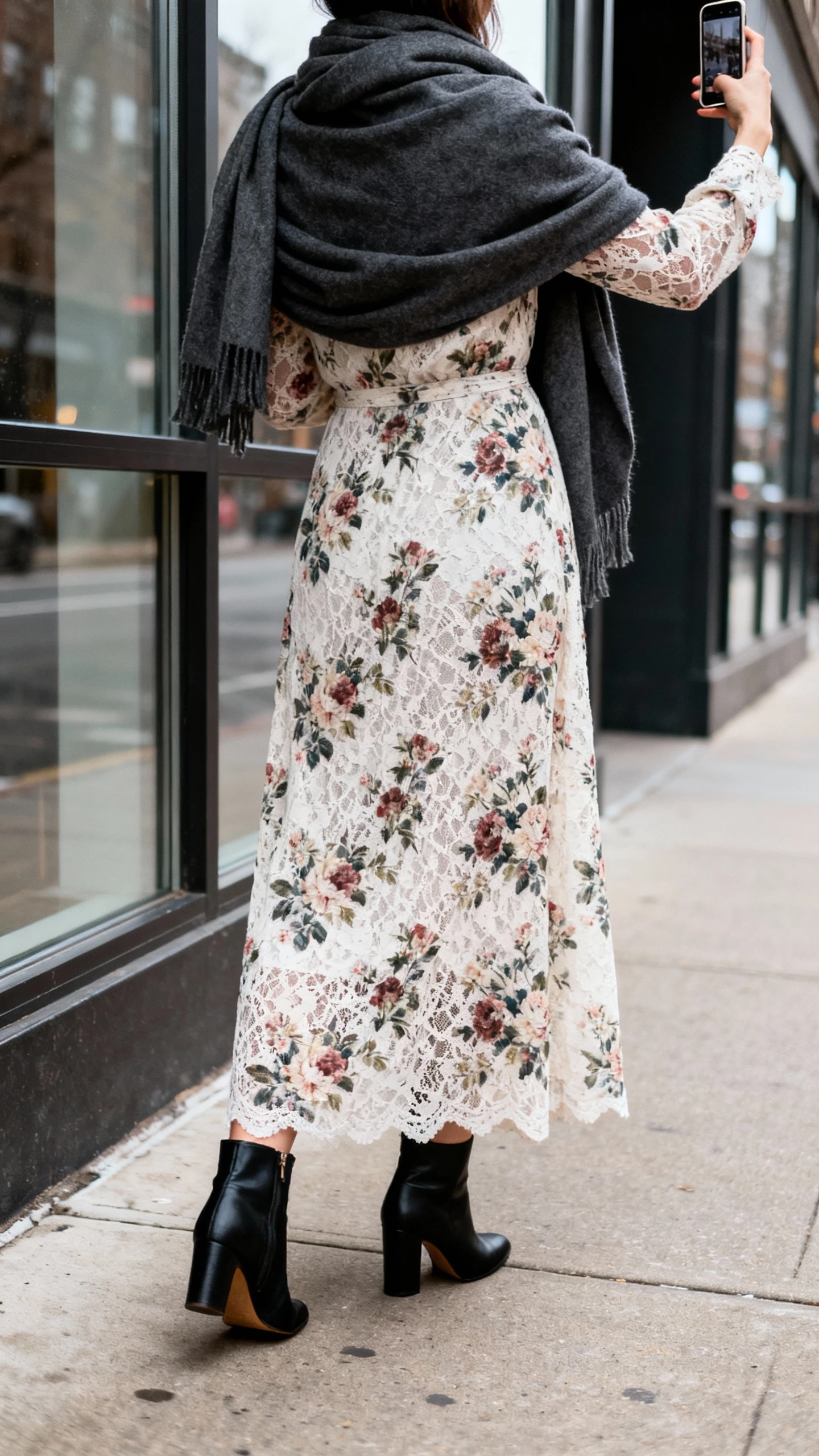 iPhone street-side back-view selfie of a woman in a floral winter-garden lace maxi dress paired with black heeled ankle boots and a charcoal wrap scarf; face not visible, city sidewalk window reflection, overcast daylight, natural iPhone aesthetic.