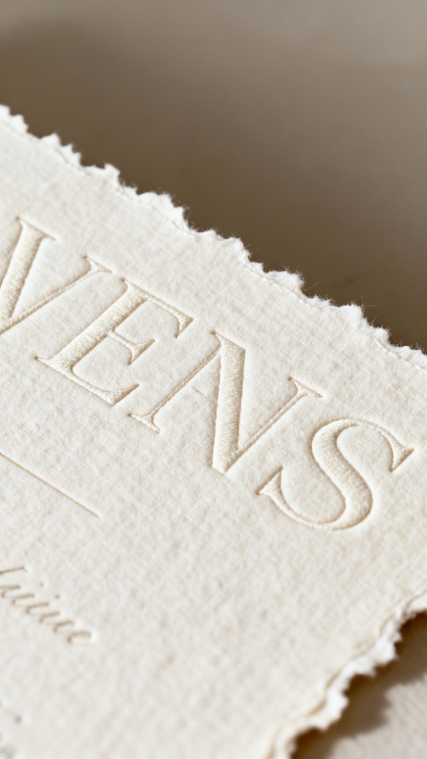 Closeup of handmade cotton invite with deckled edges, blind-embossed chunky serif, soft off-white te