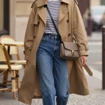 Golden-hour street style photo in a chic Parisian-inspired city setting: a confident woman mid-stride on a cobblestone sidewalk, wearing a cropped tan/camel trench jacket with a waist belt softly cinched, over a slim striped long-sleeve Breton tee. Pair with high-rise straight-leg medium-wash blue jeans, and pointed-toe black leather flats or sleek loafers. Add a structured taupe or black crossbody bag with a flap and clean lines, delicate small gold hoop earrings, and natural makeup with a soft red lip. Subtle wind lifts the trench hem, capturing effortless “I have my life together” energy. Background features classic stone buildings, café chairs, and soft bokeh traffic lights; muted, neutral palette with warm highlights. Shot on a 50mm lens at eye level, shallow depth of field, crisp textures in the trench and knit, editorial lighting, realistic fashion photography, no logos.