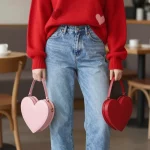 **"Fashion photography of a confident woman wearing an oversized red heart-print sweater with playful, scattered heart designs, paired with light-wash straight-leg jeans. The sweater is casually half-tucked to accentuate the waist, creating a relaxed but stylish look. She completes the outfit with clean white chunky sneakers and holds a mini heart-shaped bag in soft pink or red for a romantic touch. The background is a cozy, neutral-toned café with warm lighting, perfect for a Galentine's brunch or casual date. Soft natural light enhances the textures of the knit sweater and denim, capturing a candid yet chic Valentine’s Day aesthetic. The composition is balanced and aspirational, with a focus on effortless romance."**