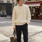 **Image Prompt:** A stylish, effortlessly chic man with tousled hair stands on a cobblestone Parisian street, exuding romantic European charm. He wears a slightly oversized cream cable-knit sweater, casually tucked in at the front, paired with dark tailored trousers cuffed once to reveal sleek tan leather sneakers. A delicate silver chain necklace adds subtle elegance, while a structured tote bag rests casually by his side, hinting at a poetic, bookish vibe. Soft morning light filters through the trees, casting a warm glow on the scene, capturing the perfect balance of coziness and sophistication. The background features classic Parisian architecture with a cozy café nearby, enhancing the romantic, timeless aesthetic. The composition is styled like a high-fashion editorial—natural, candid, and effortlessly polished.