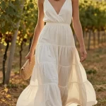A stunning, high-fashion photo of a woman wearing a milk-white tiered cotton midi dress that flows gracefully as she walks, the delicate layers catching the soft light with a subtle textured elegance. She pairs it with braided leather sandals, a wooden bead necklace, and a woven clutch in natural tones, embodying minimalist romance. Her slightly wavy hair and minimal makeup enhance the undone, earthy vibe, while the wooden beads add a warm contrast. The scene is set at a sunlit vineyard wedding, with golden hour light casting a dreamy glow, emphasizing the dress's liquid-like movement and effortless beauty. The composition is editorial yet natural, focusing on the dress's flow and the serene, boho-chic aesthetic.